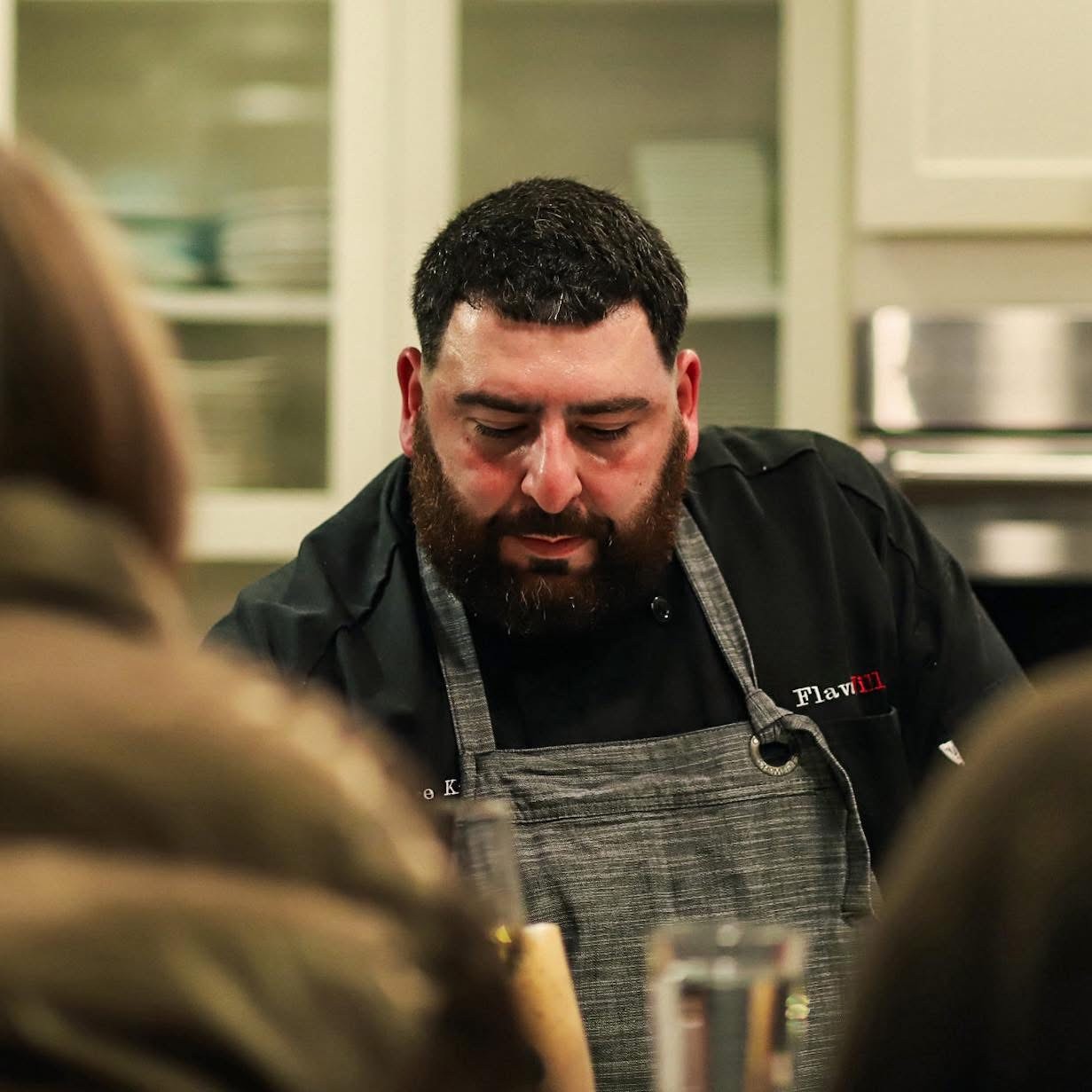 Bearded chef in black jacket and apron, head down, working.