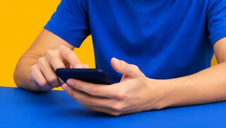 Close-up of hands using a smartphone on a blue table against a bright yellow background.