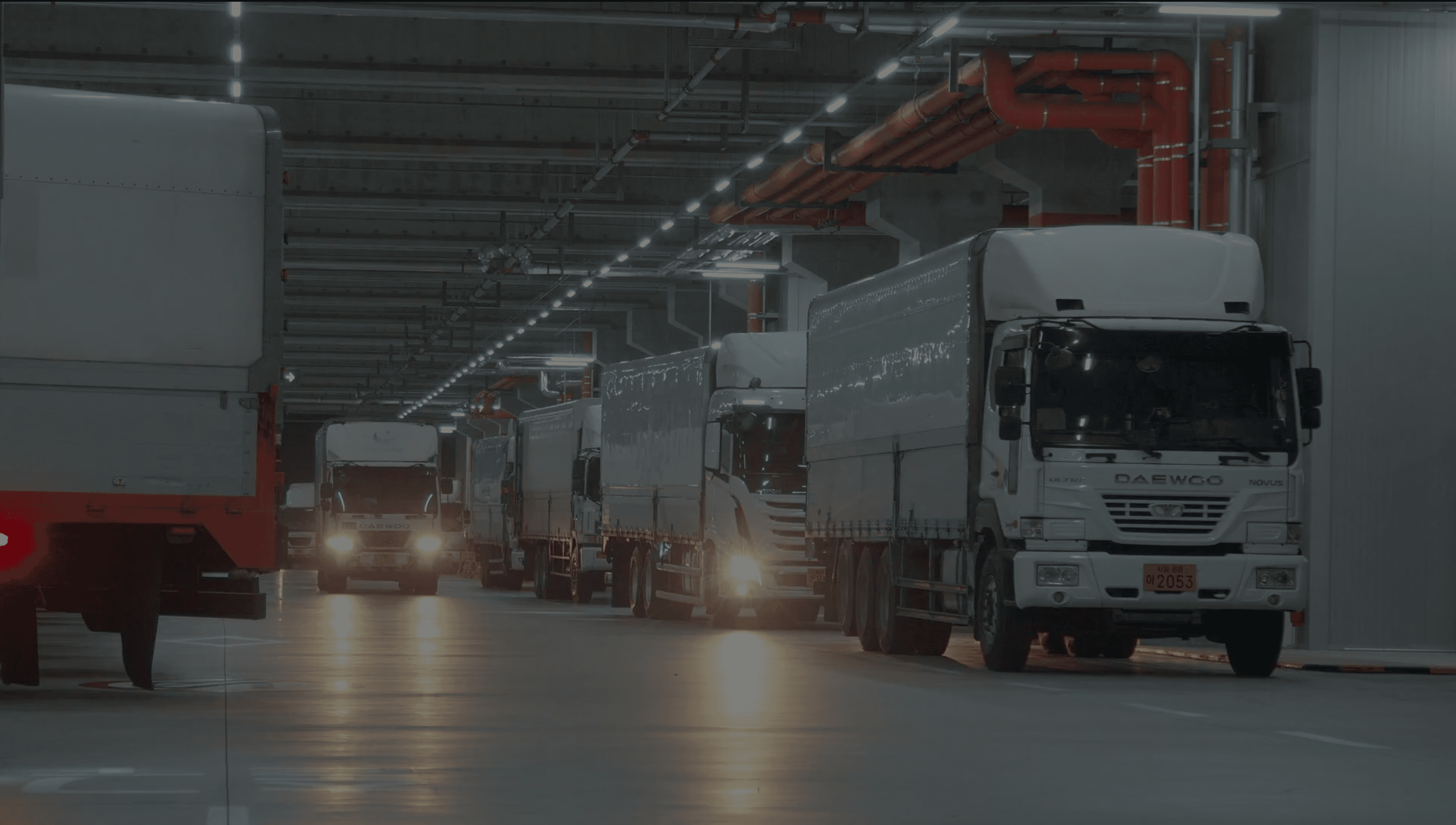 CJ대한통운 더 운반 소속 대형 화물 운송 차량들이 실내 물류센터에 일렬로 정차해 있는 모습입니다. 전국 네트워크를 갖춘 운송 차량과 인프라를 통해 합리적인 운임으로 안정적인 배차 서비스를 제공함을 보여줍니다.