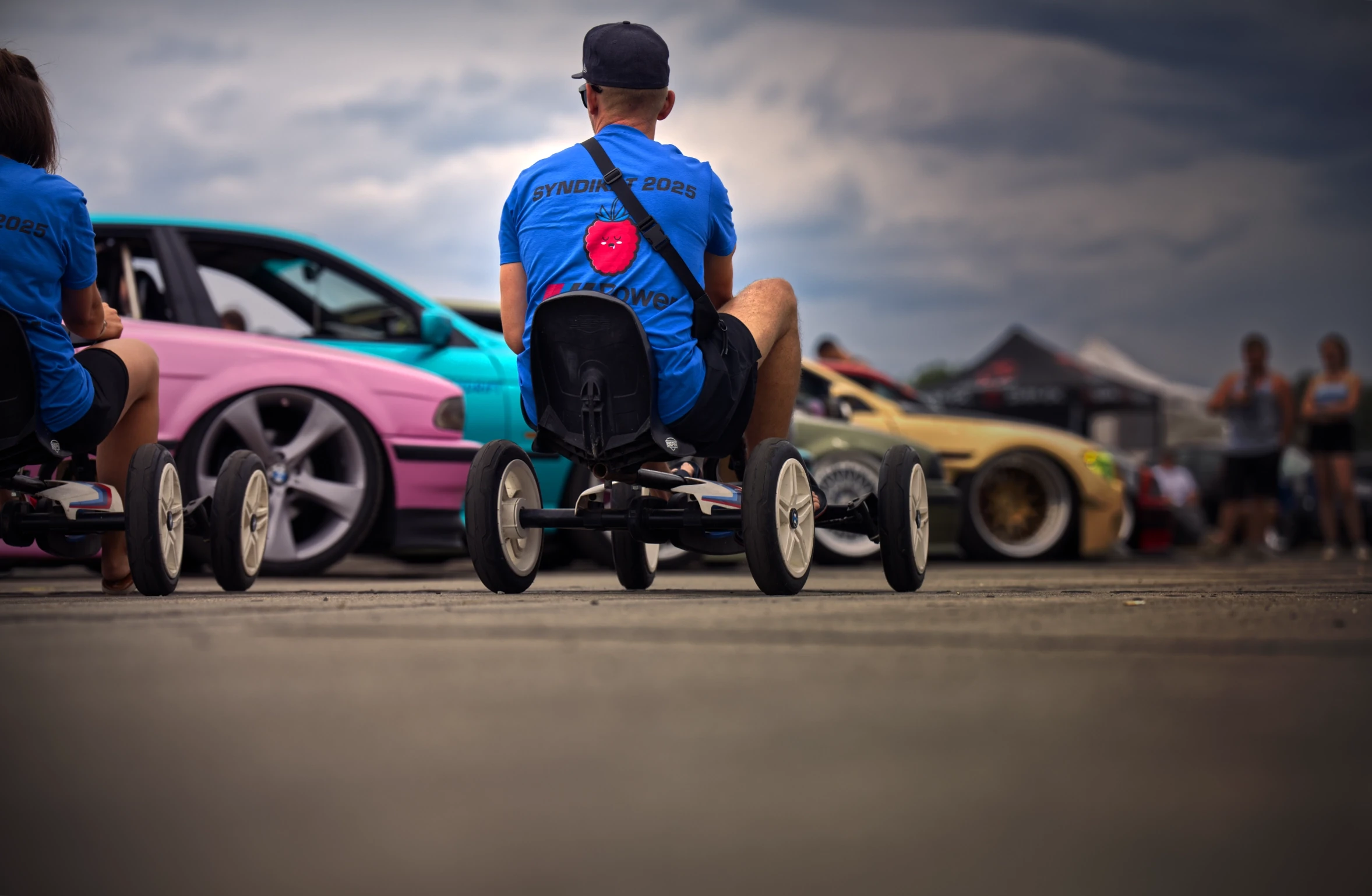 Besucher auf Gokarts vor individuell gestalteten BMW Fahrzeugen beim BMW Syndikat Asphaltfieber 2025 unter dramatischem Himmel.