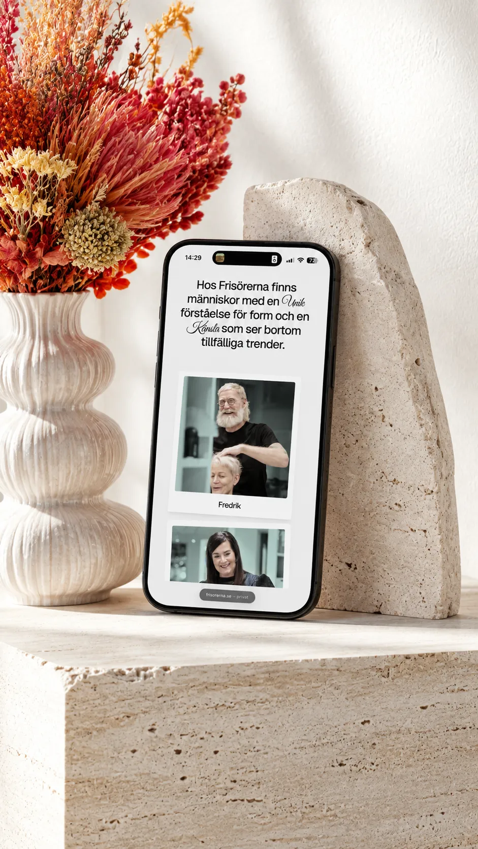 iPhone displaying the Frisörerna team page, supported by a sculptural stone object beside dried flowers.