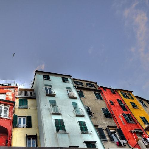 Barevné budovy s balkony proti modré obloze, s červenými, žlutými, modrými a béžovými fasádami. Na obloze letí pták.