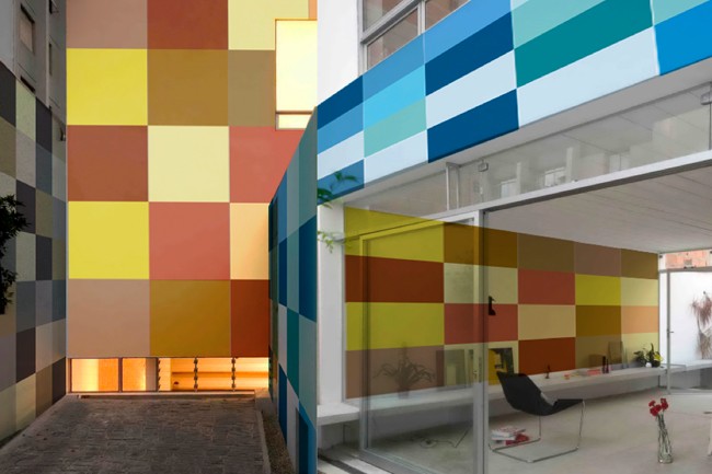 A modern building exterior features a colorful, geometric wall composed of square panels in shades of yellow, orange, brown, and teal. A glass sliding door reveals a room interior with a black chair and a vase of red flowers. A paved walkway runs alongside the wall.