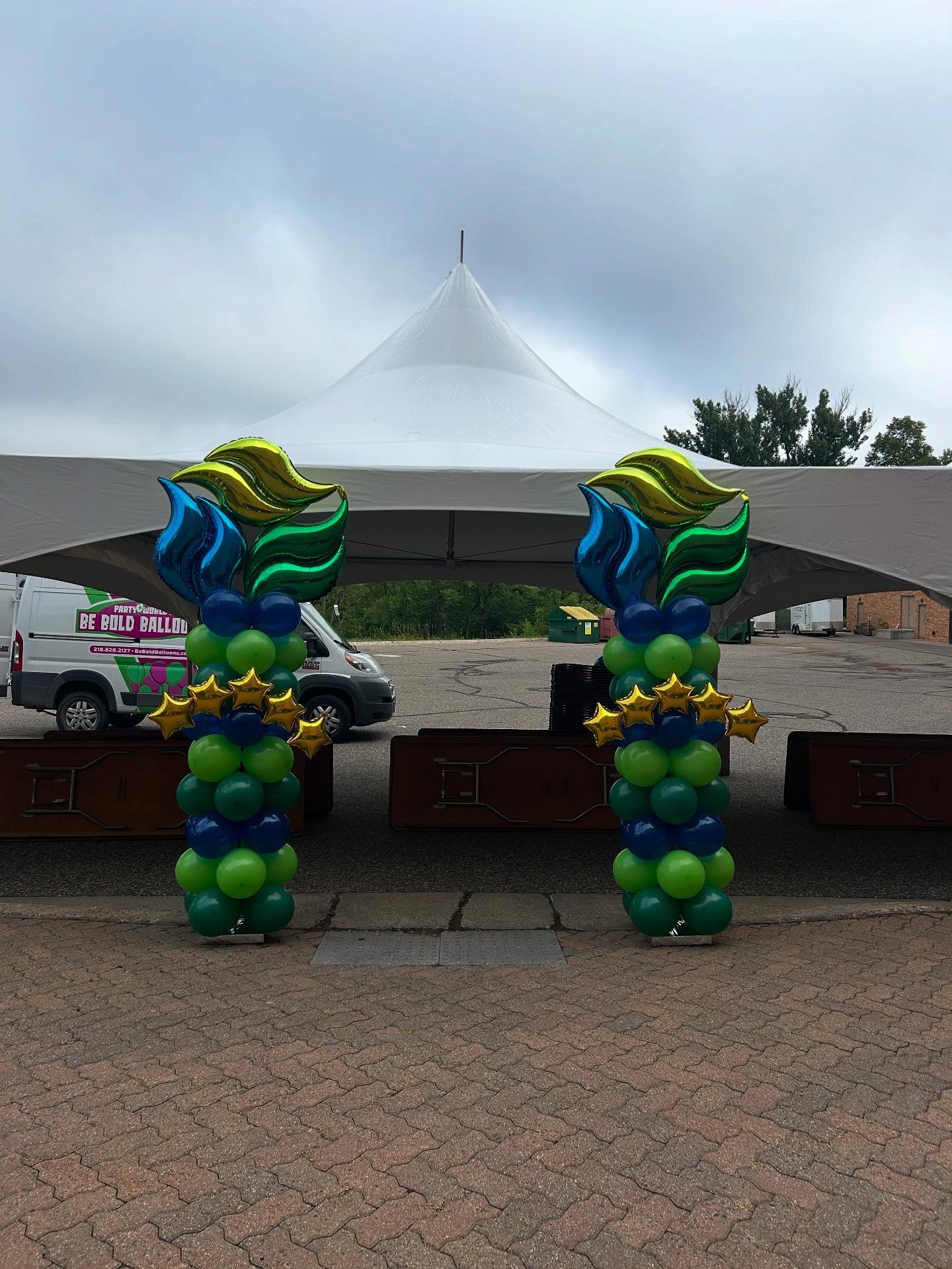 Green, blue, and yellow balloon columns with starburst toppers arranged outdoors near a white tent by Be Bold Balloons