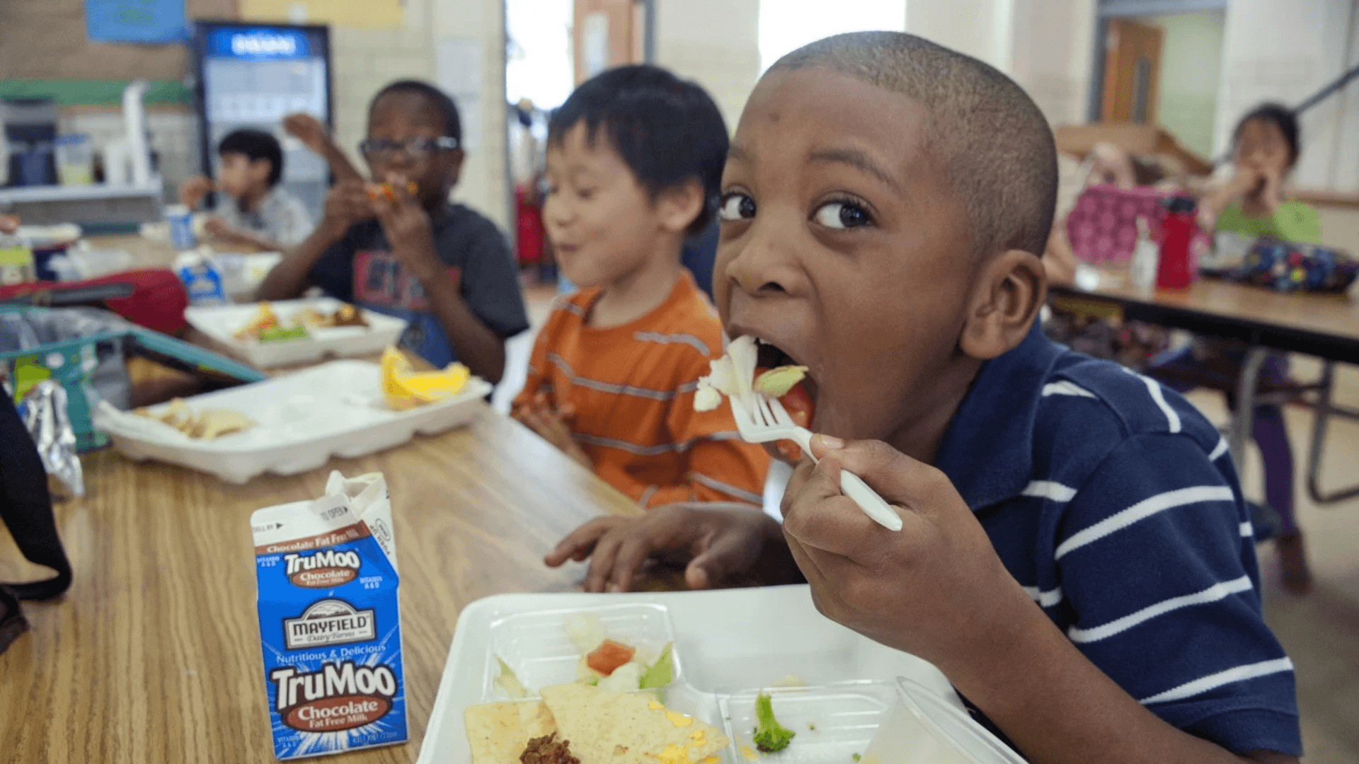 Elementary school students eating lunch in a cafeteria, with a boy in the foreground taking a bite of salad while seated at a table with classmates.