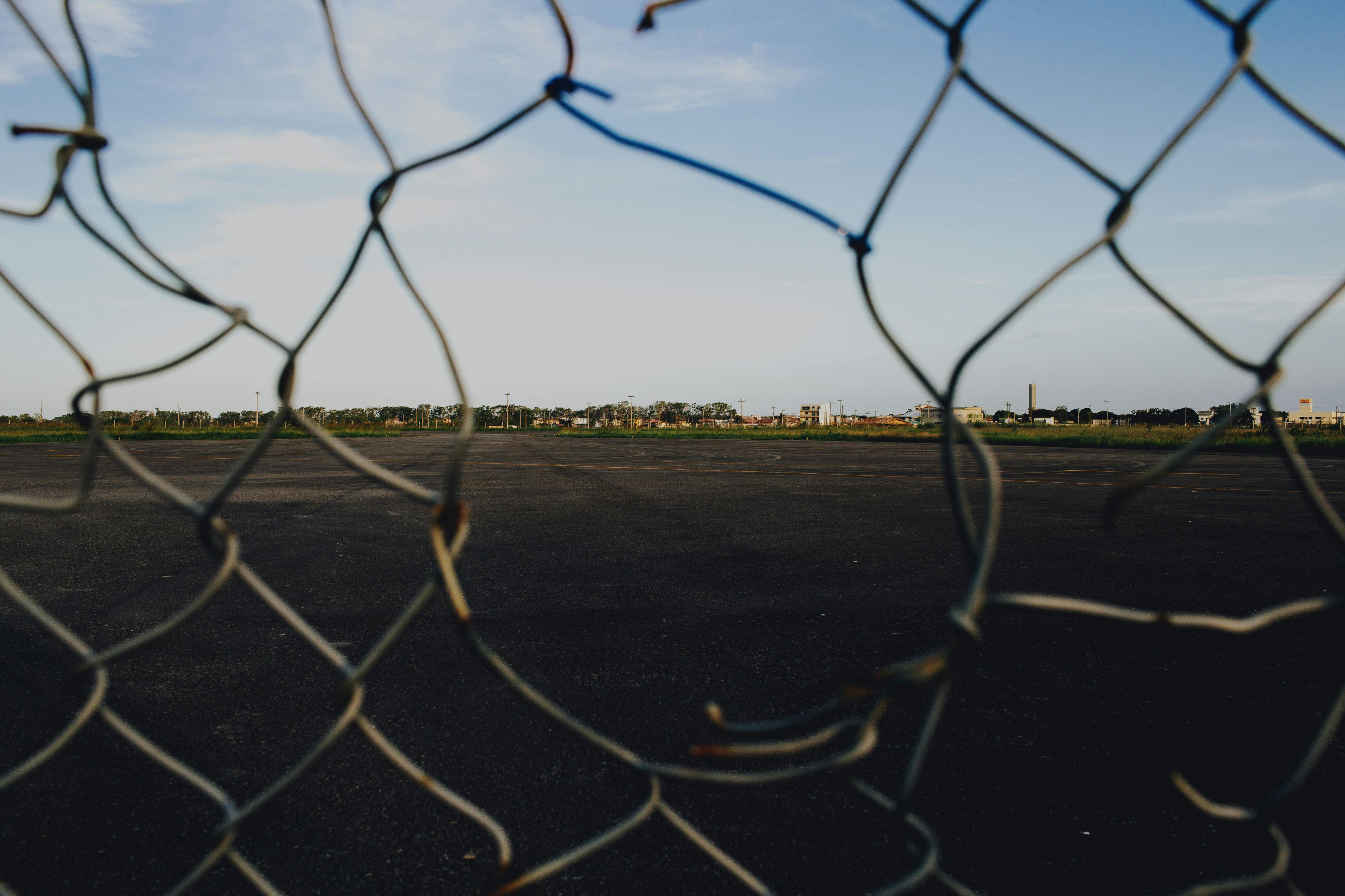 A broken wire mesh fence framing an open asphalt area under a blue sky.