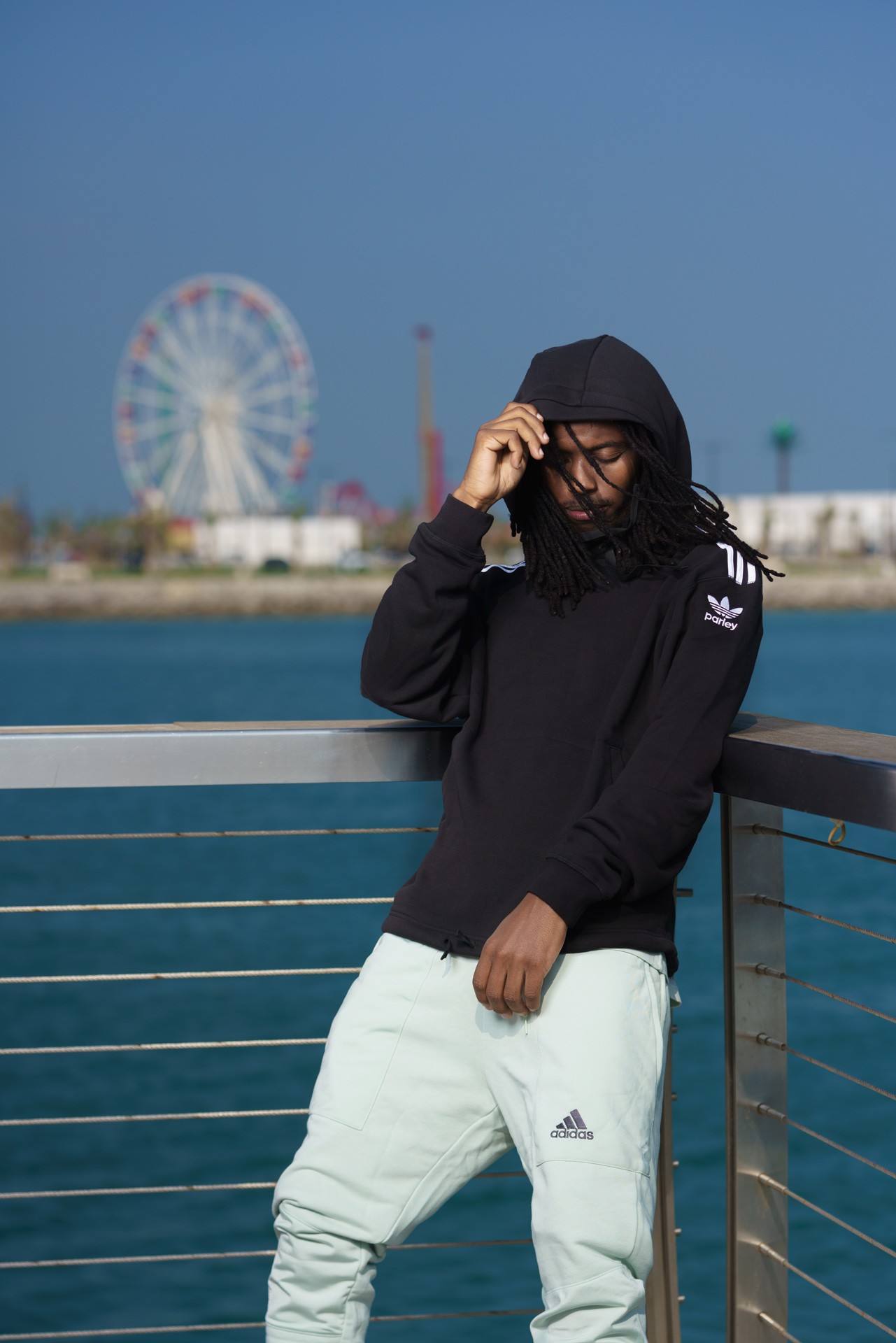 A man in a black adidas hoodie and mint joggers leaning on a railing by the water, a Ferris wheel behind him.