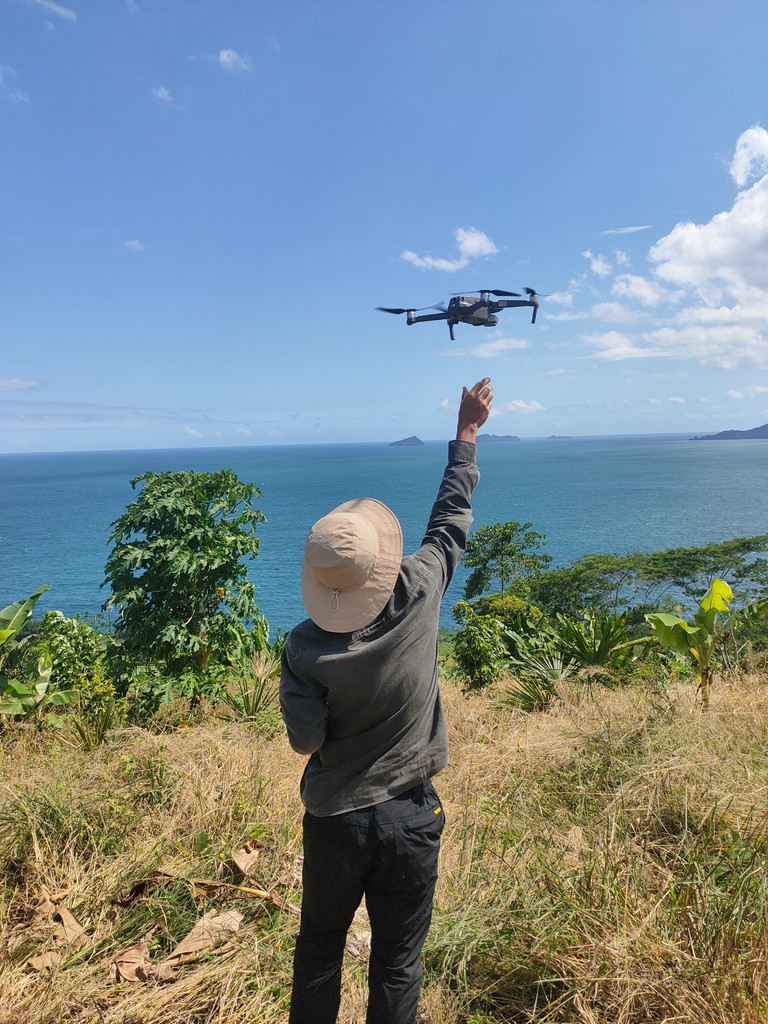 Person holding a drone above them, with a scenic view of the ocean and blue sky in the background.