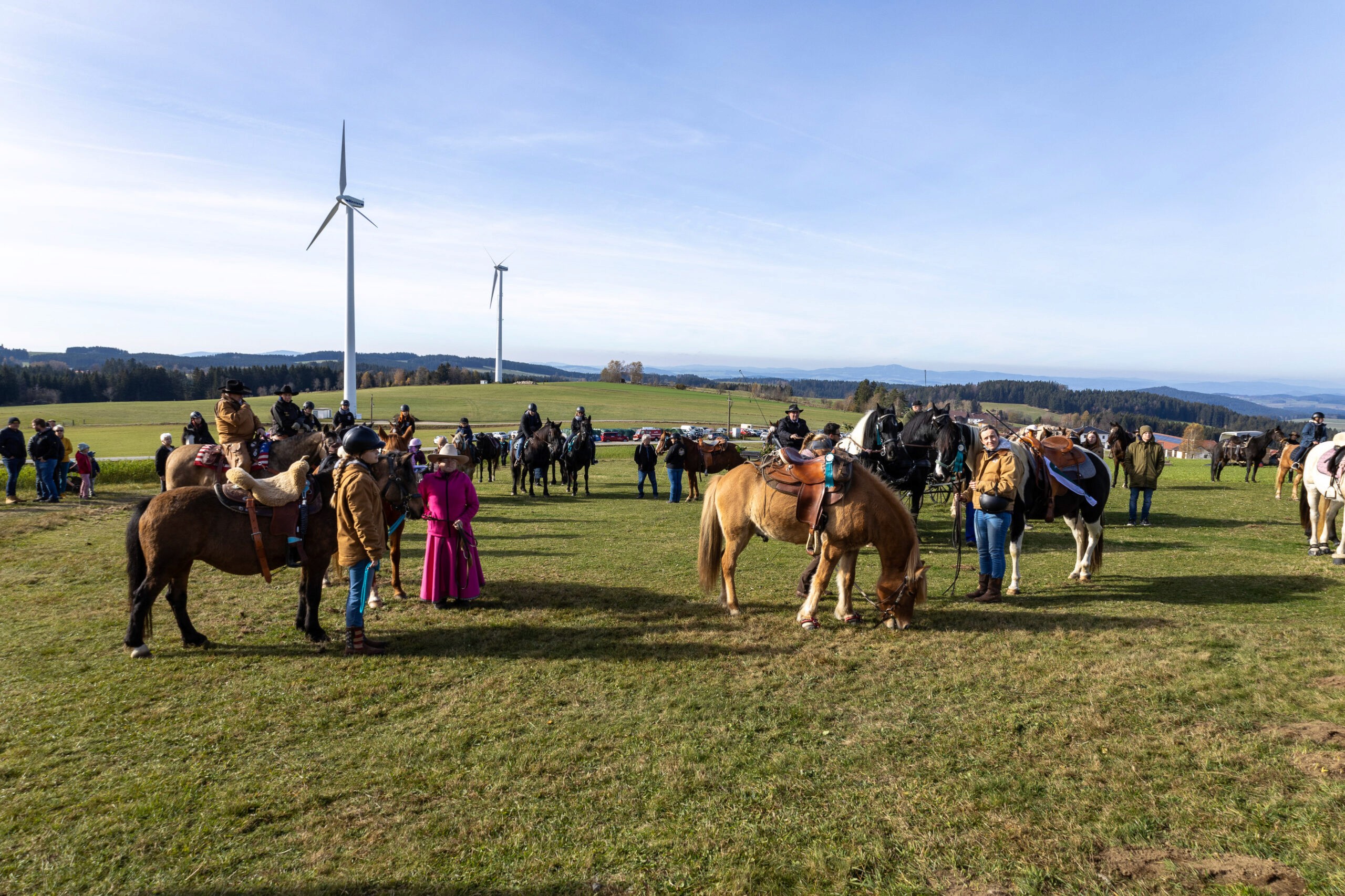 Pferde in Wanderreitausrüstung grasen auf einer Wiese neben ihren Reitern im Hochland. Im Hintergrund ein weiter Ausblick und Windmühlen.