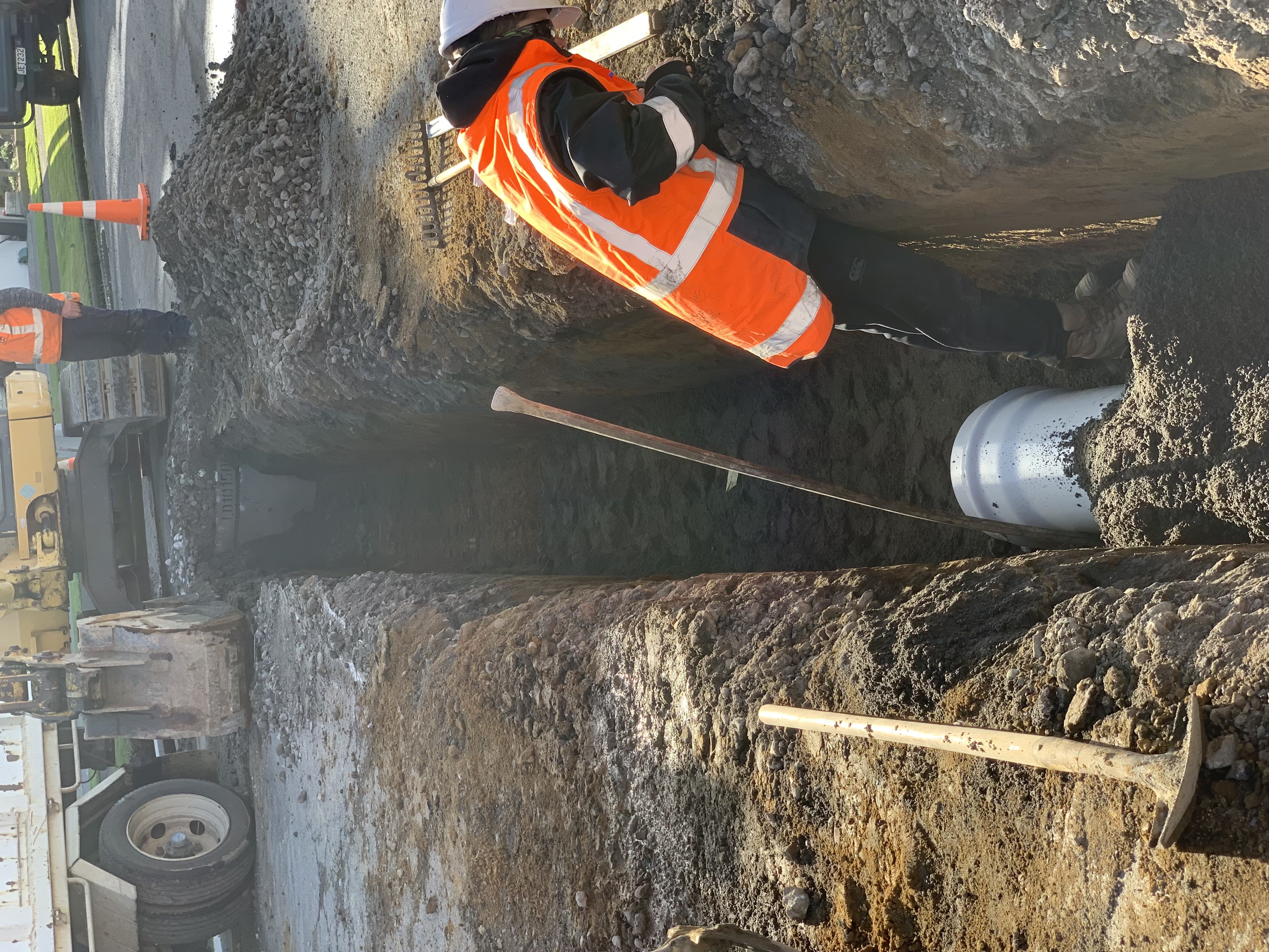 Worker standing beside a pipe connection in a narrow excavated trench.