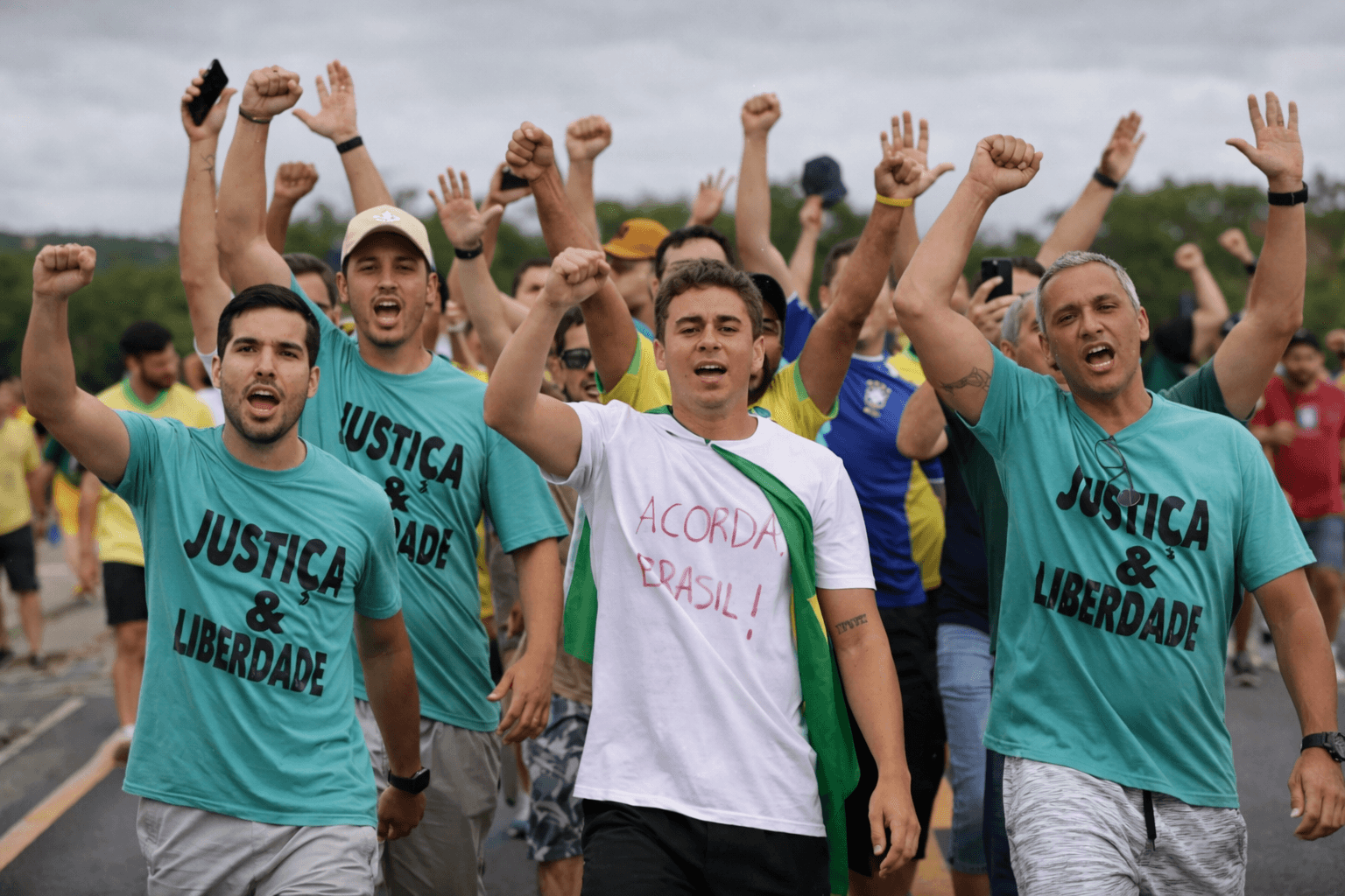 Multidão de pessoas caminhando juntas em manifestação ao ar livre, com braços erguidos e punhos cerrados, vestindo camisetas com frases como “Justiça & Liberdade” e “Acorda Brasil”, em uma estrada sob céu nublado.