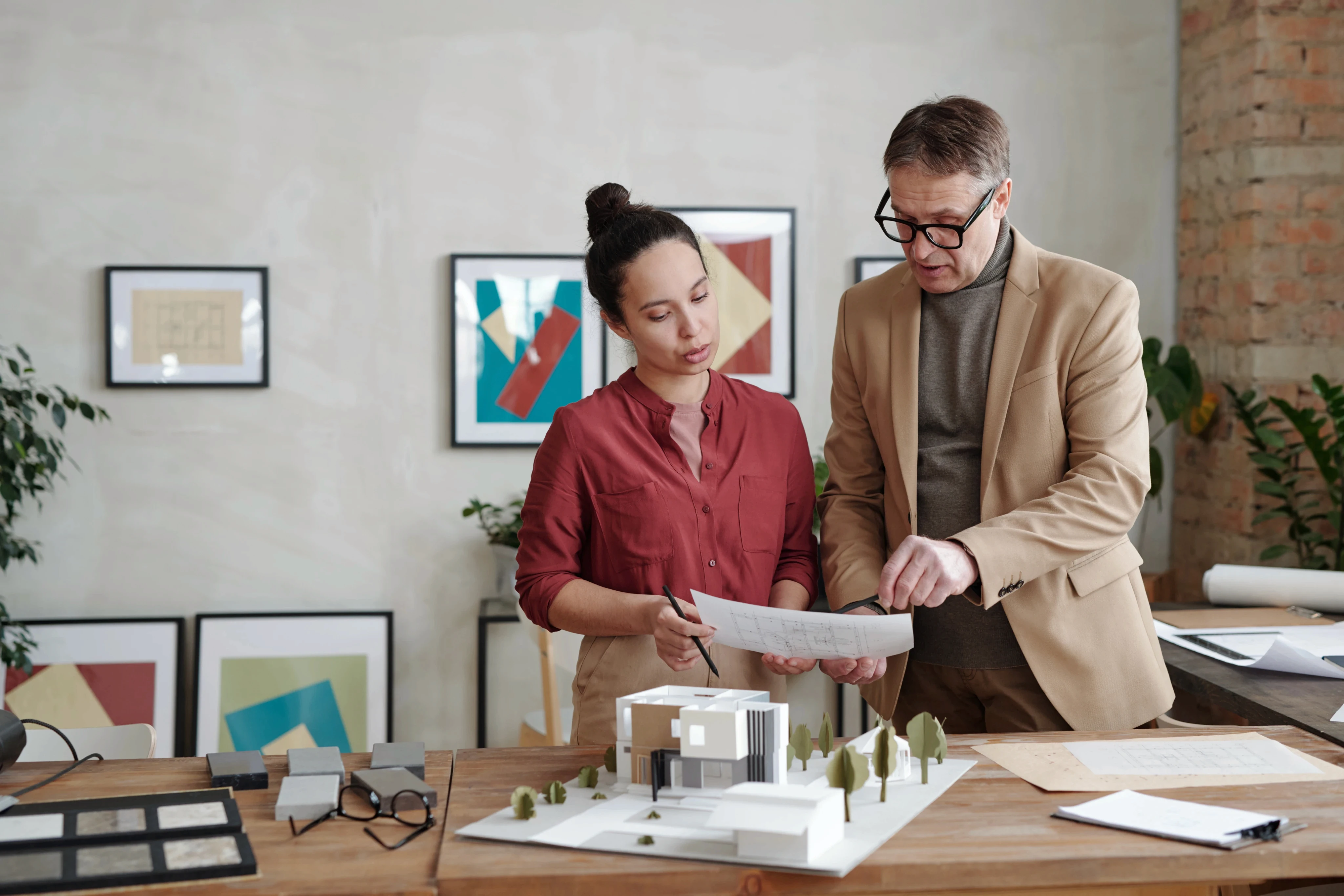 A man and a woman review architectural plans beside a building model on a desk. Art frames hang on a wall behind them, adding a creative atmosphere.