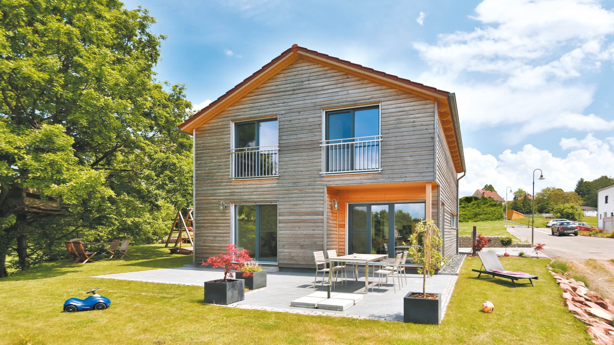 Modernes Holzfertighaus mit Terrasse, Gartenmöbeln, Spielzeug und viel Grün bei sonnigem Wetter.