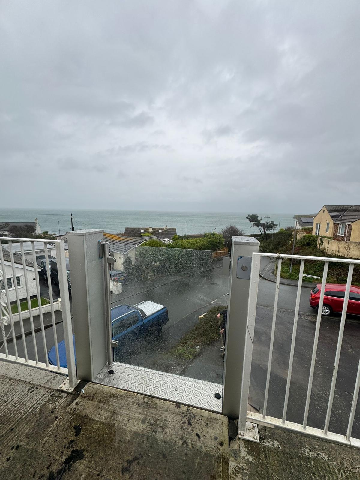 View from upper balcony through open vertical platform lift enclosure at a coastal UK property — sea view beyond with residential street and parked vehicles below