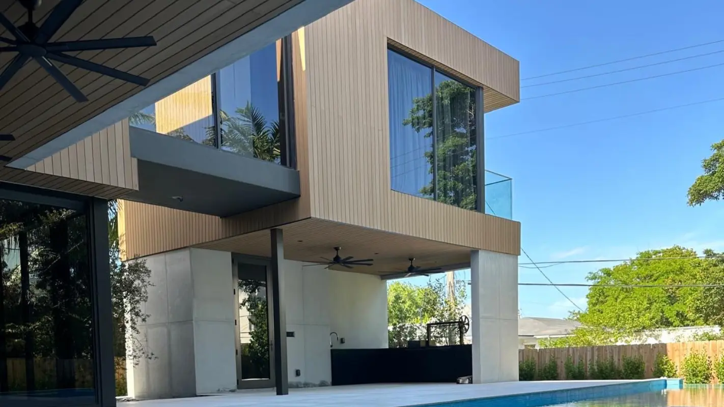 Backyard view of Meridian Avenue Residence in South Beach — covered outdoor living area with ceiling fans and summer kitchen beneath a cantilevered wood-clad upper level, with infinity pool by Atelier International.