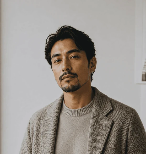 Man with dark hair & beard, neutral expression, wearing light brown sweater & jacket. Light background.