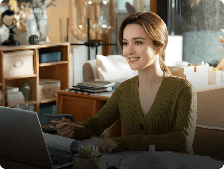 Company name check - Smiling woman working on laptop, holding pencil, looking at screen, wearing green sweater.