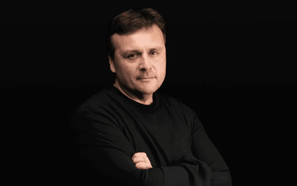 A person with short, dark hair, wearing a black long-sleeve shirt, stands against a dark background with their arms crossed and a neutral expression.