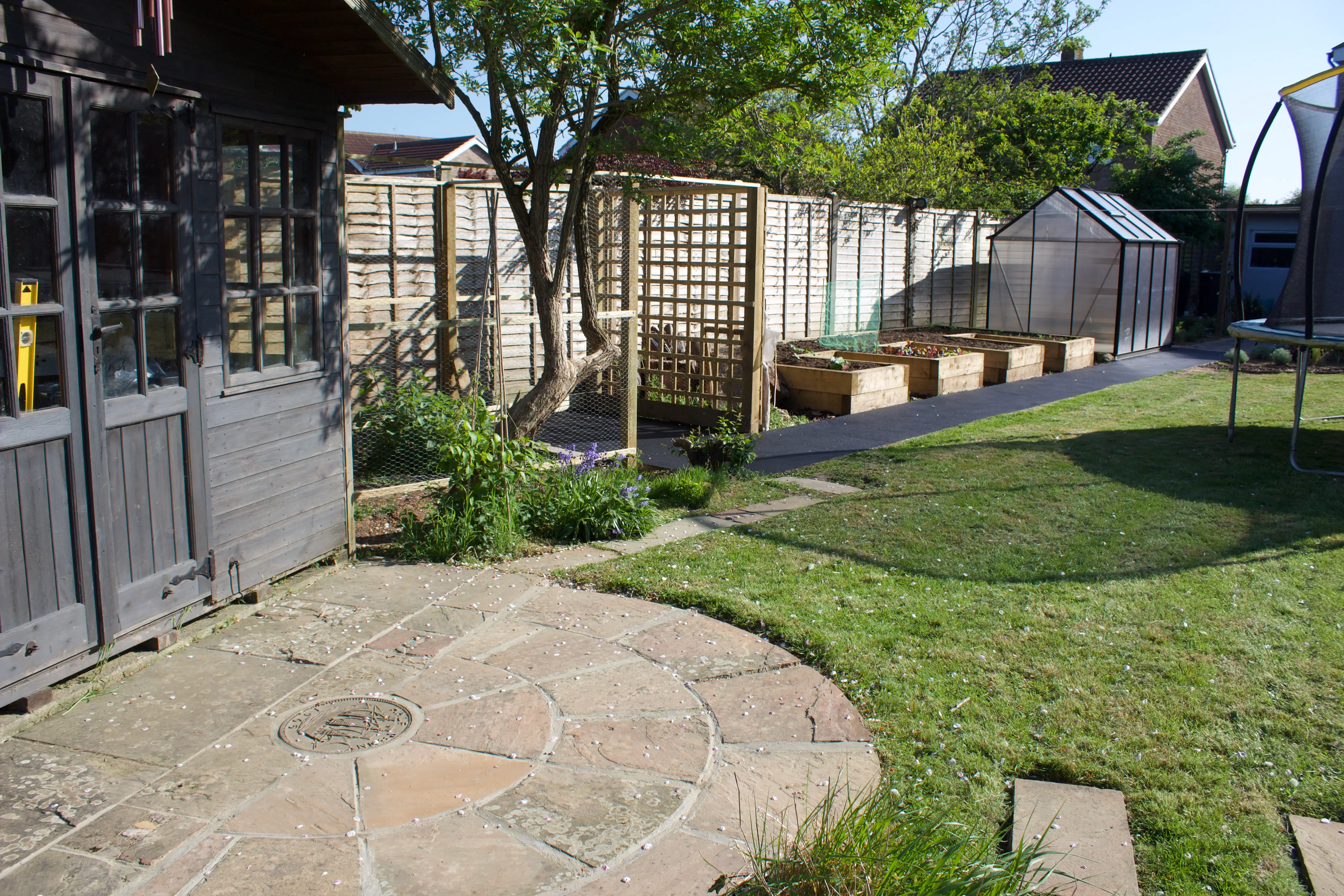 A sunny garden featuring a curved stone path, green grass, and neatly arranged shrubs beside wooden fences.