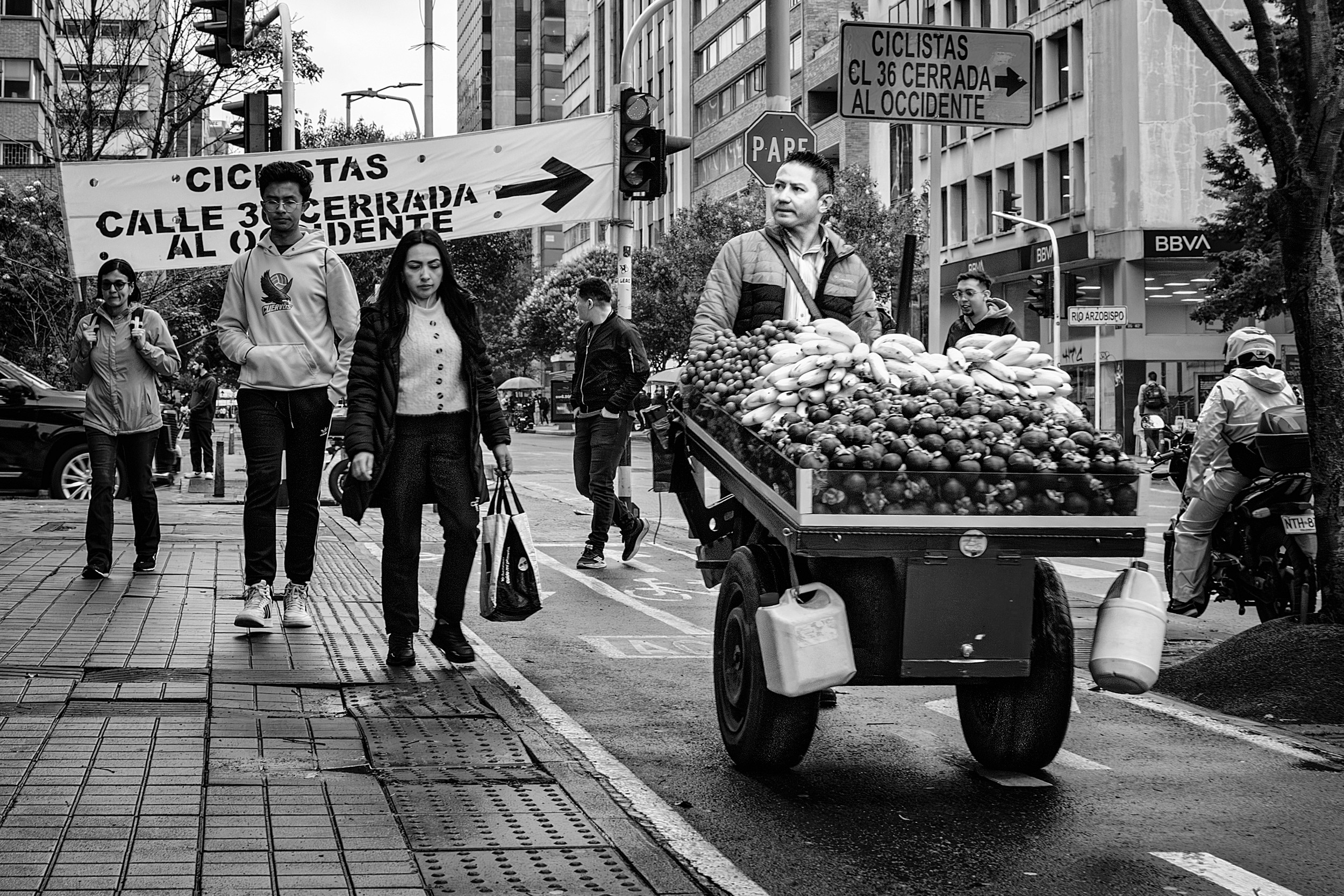 A street vendor pushes a cart overflowing with bananas and produce through a busy Bogotá intersection, surrounded by pedestrians and city signs.