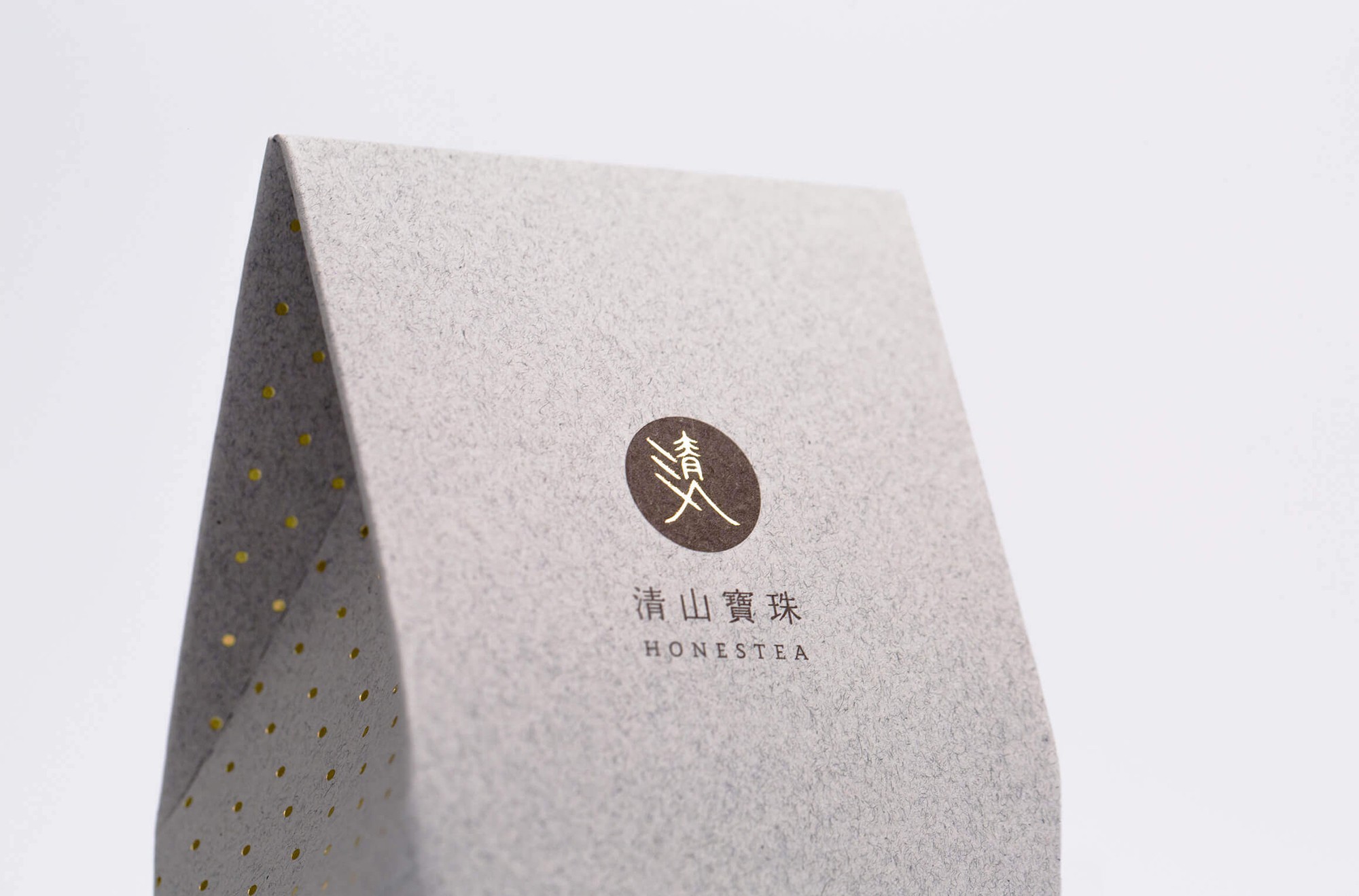 這張圖展示了清山寶珠的包裝設計。包裝使用灰色紙材質，正面印有清山寶珠的標誌和品牌名稱「HONESTEA」，標誌為圓形金色圖案，裡面帶有黑色抽象的中文字「清山」。