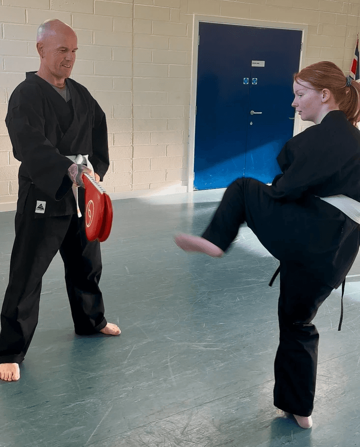 A student holds a kick pad as another student practices a kick. The setting is a brightly lit training room.