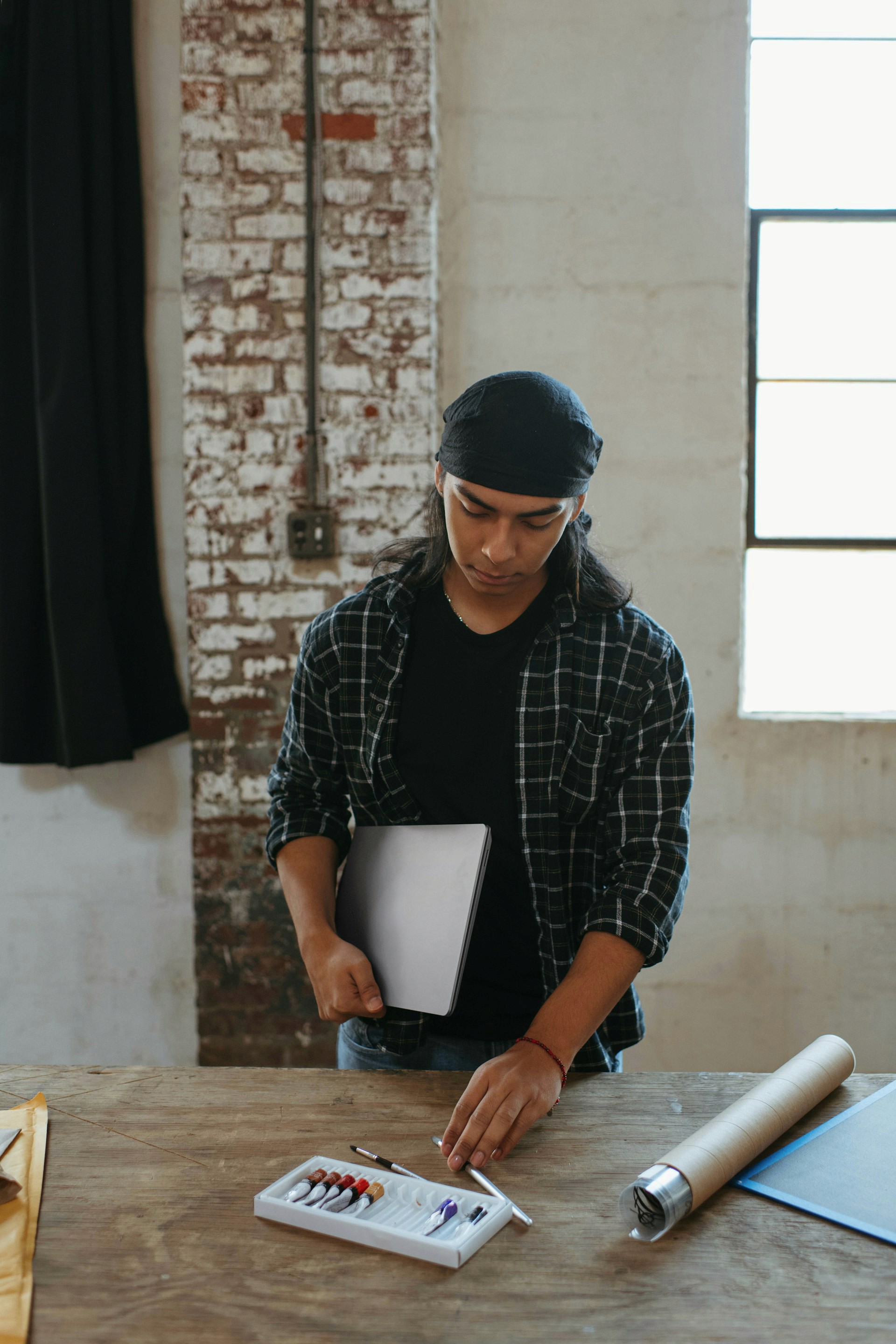 Young person wearing a bandana and plaid shirt holding a silver laptop next to a wooden table with tools.