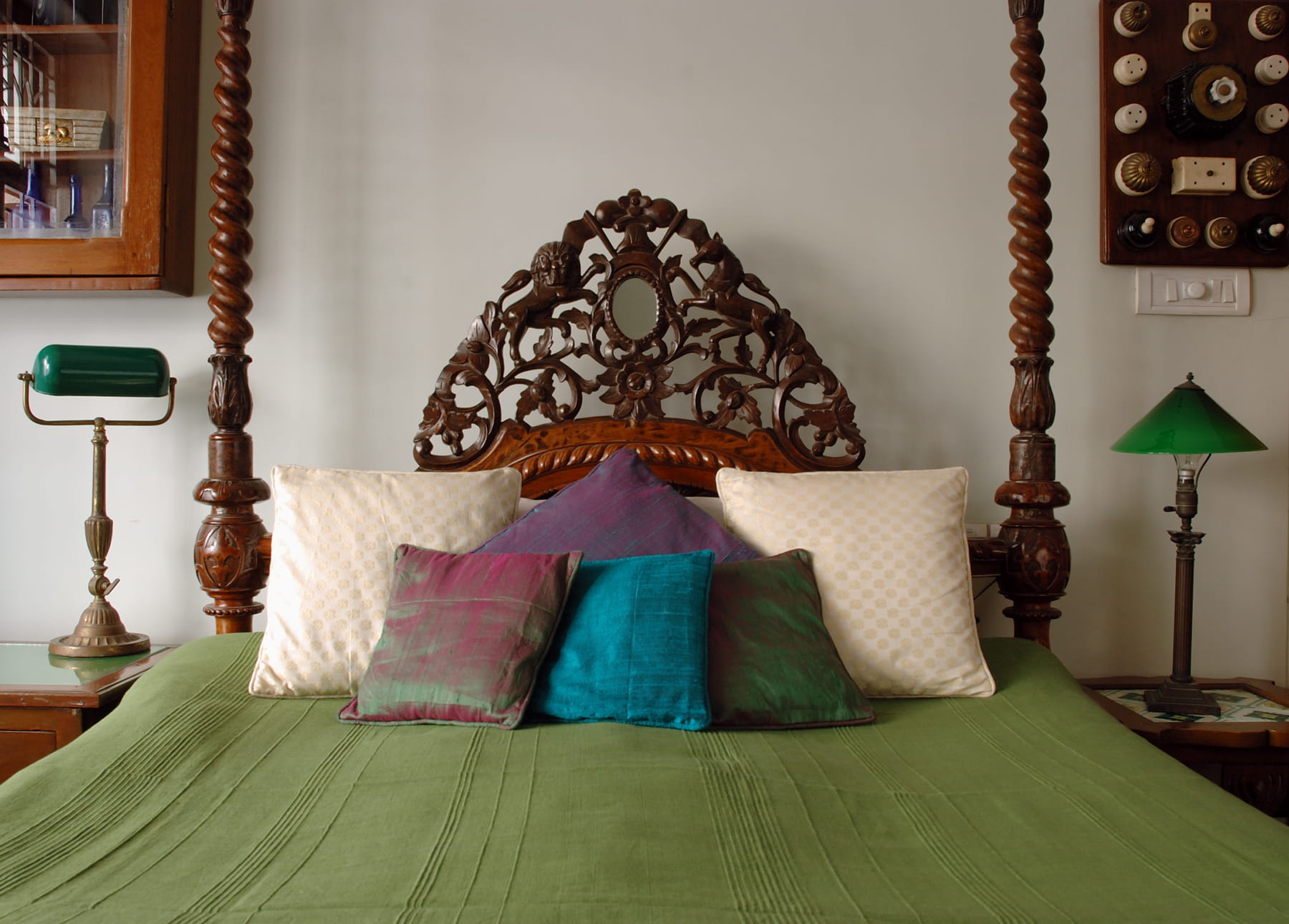 Carved wooden four-poster bed with green bedding and layered jewel-toned cushions.