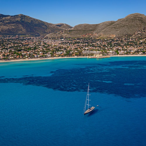 A sailboat on clear blue water near a coastline with buildings and mountains in the background under a clear sky.