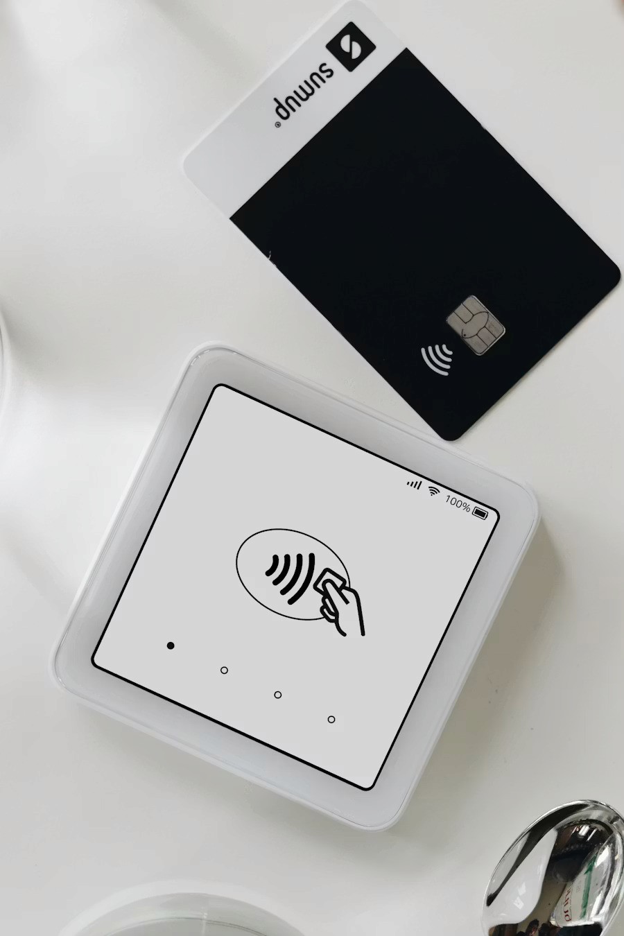 A contactless payment terminal with a sleek, minimalistic design is shown on a white surface, along with a black credit card positioned above it.