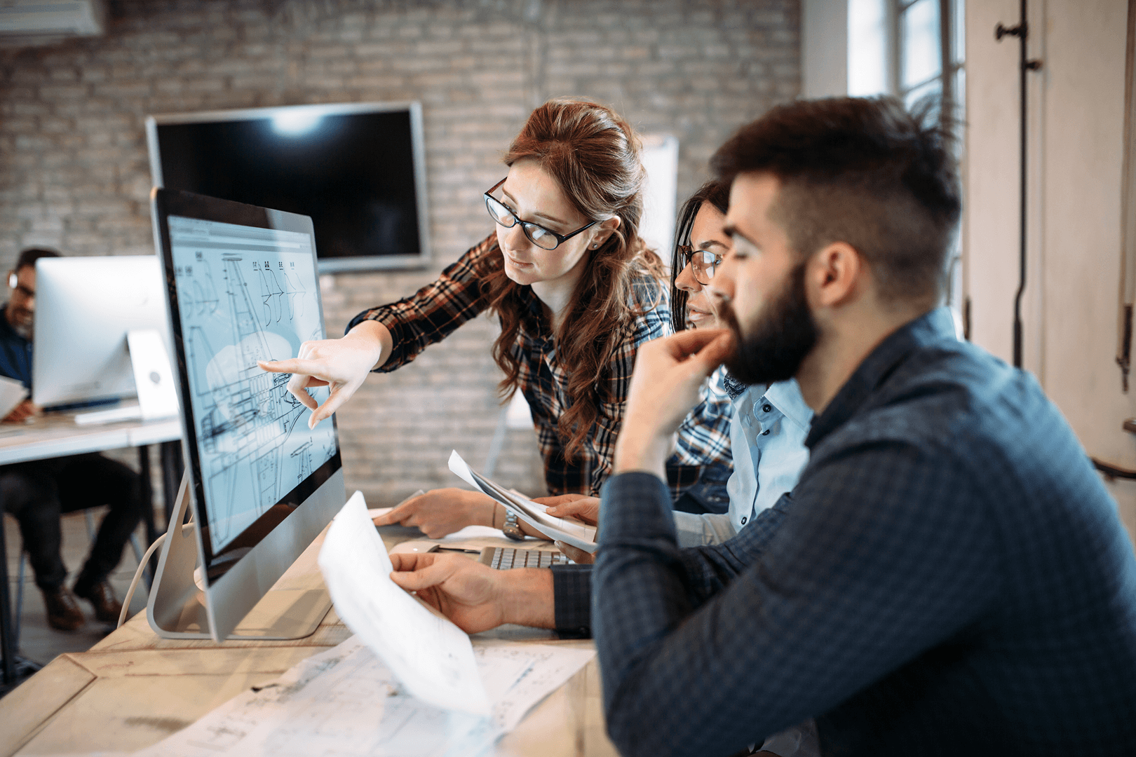 Woman pointing at technical drawings on a computer while discussing with colleagues