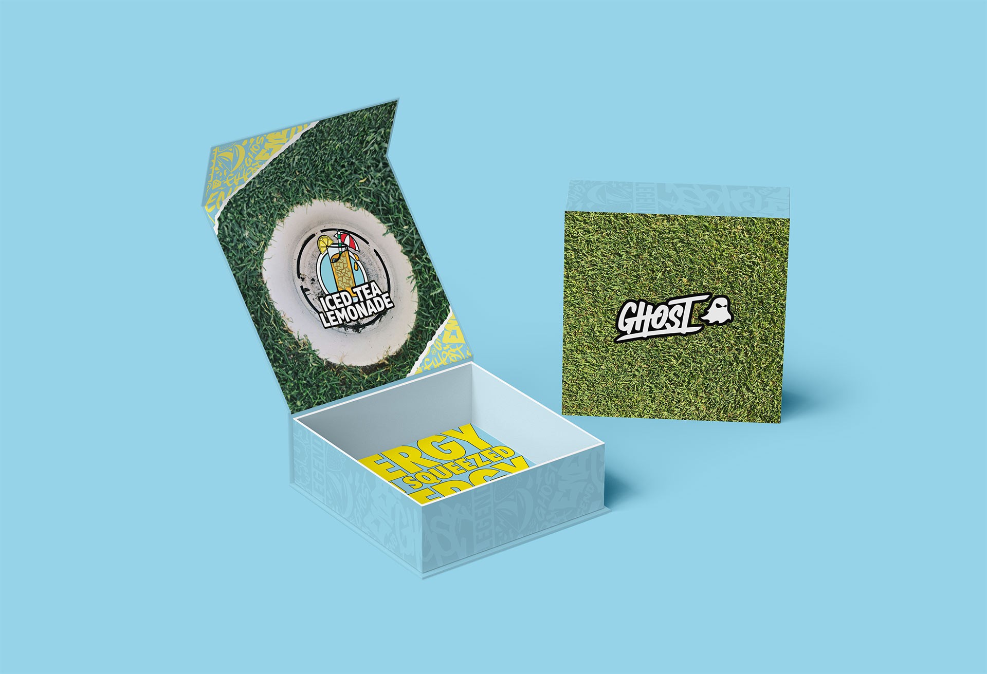 Promotional packaging mockup for a Ghost campaign featuring a custom box design with grass-textured graphics, bright yellow typography, and branded iced tea lemonade visuals against a light blue background.