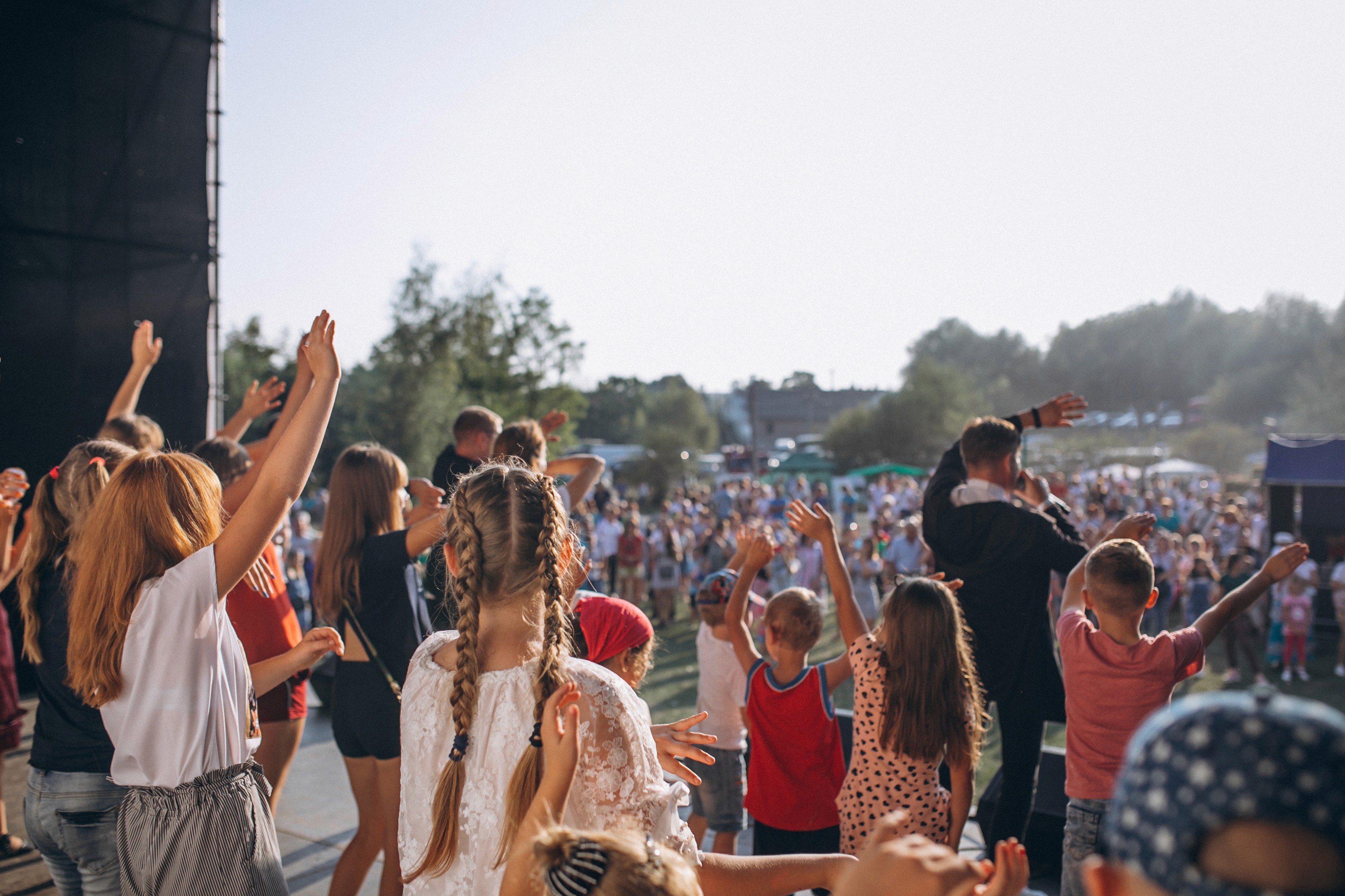 Enfants et animateur sur scène levant les bras devant une foule lors d’un festival en plein air.