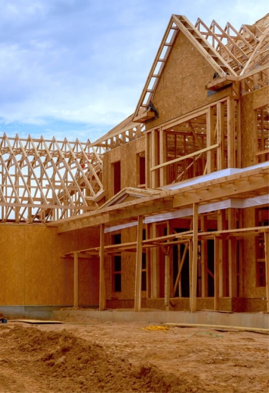 A partially constructed wooden house with a sandy foundation and framing visible under a clear blue sky.
