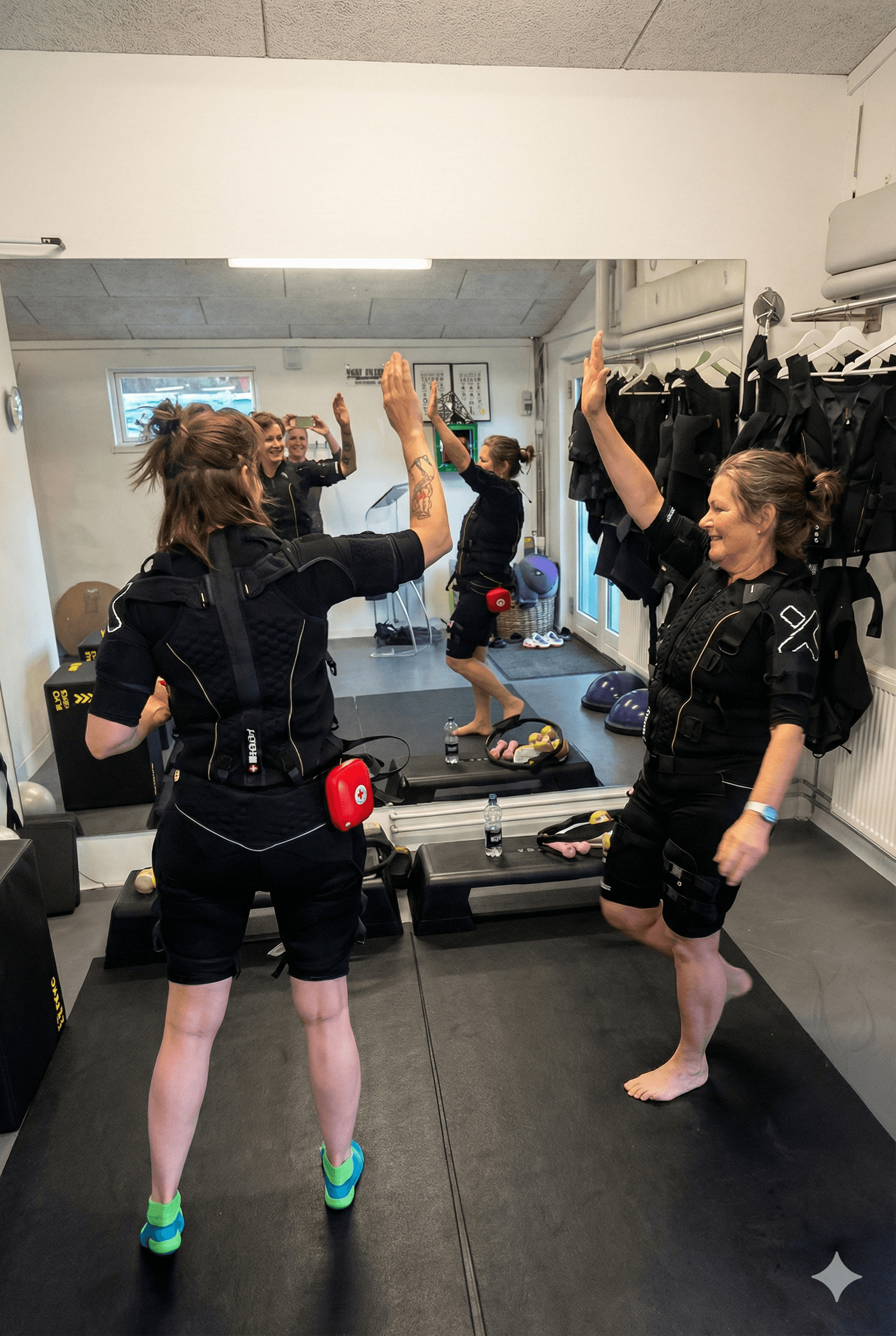 A muscular man smiling while performing a hanging leg raise on gymnastic rings in a bright fitness studio.