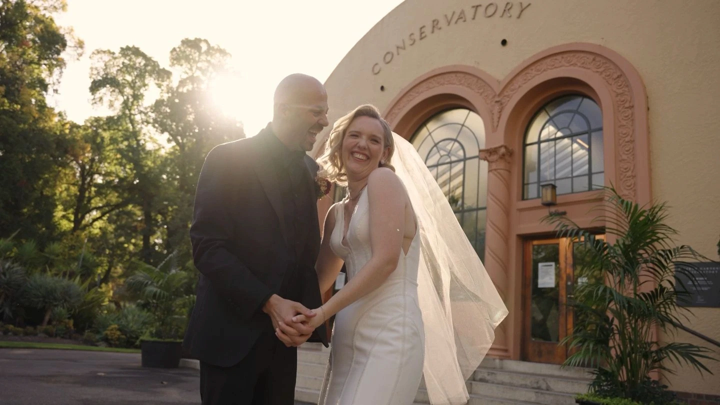 A couple in wedding attire joyfully holds hands outside an elegant conservatory building, with the sun softly illuminating the scene and lush greenery surrounding them.