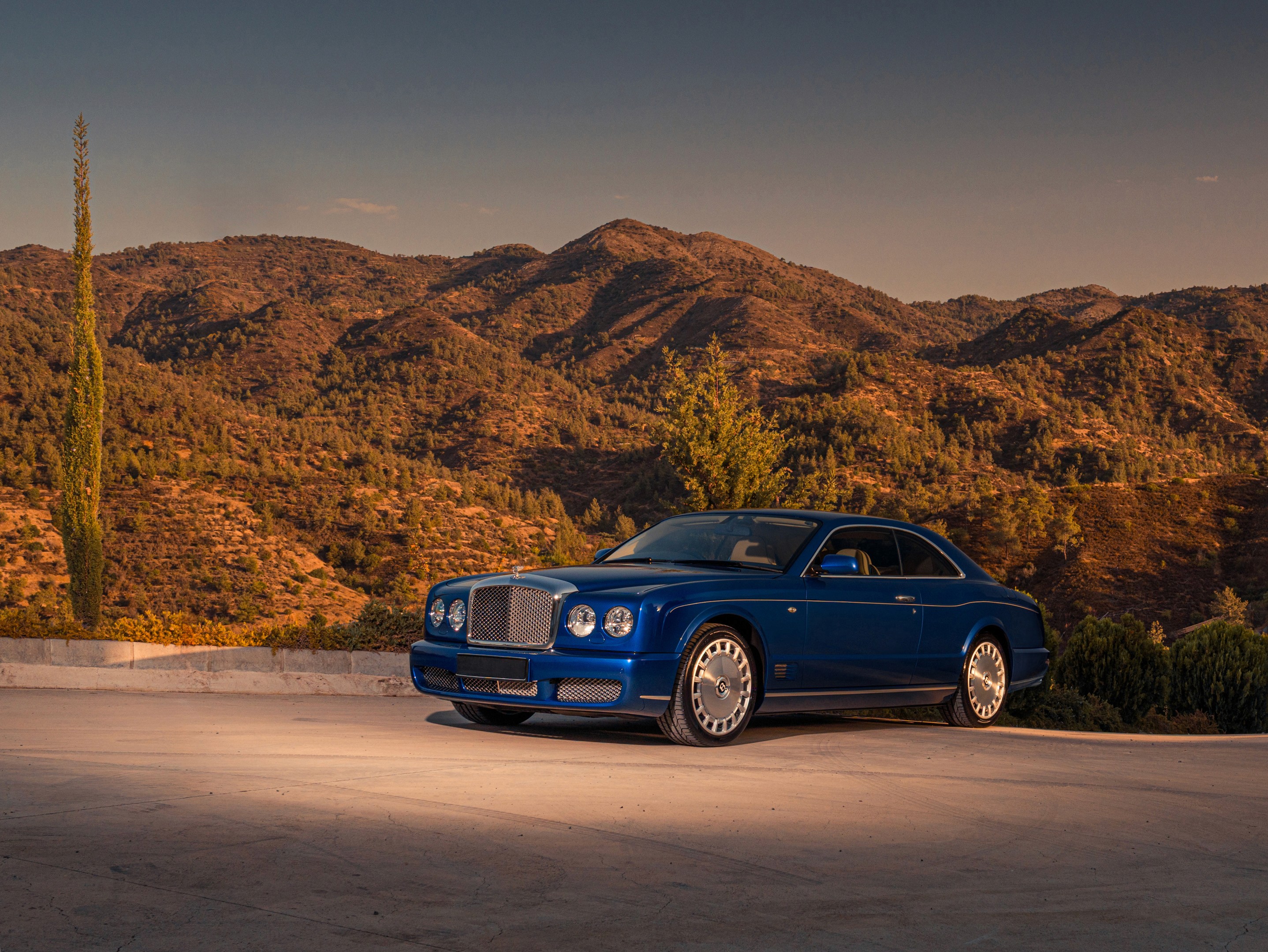Bentley Brooklands coupé in Moroccan Blue photographed from the front three-quarter, chrome mesh grille and quad round headlamps facing forward, against the tree-covered foothills of Troodos, Cyprus in warm evening light.