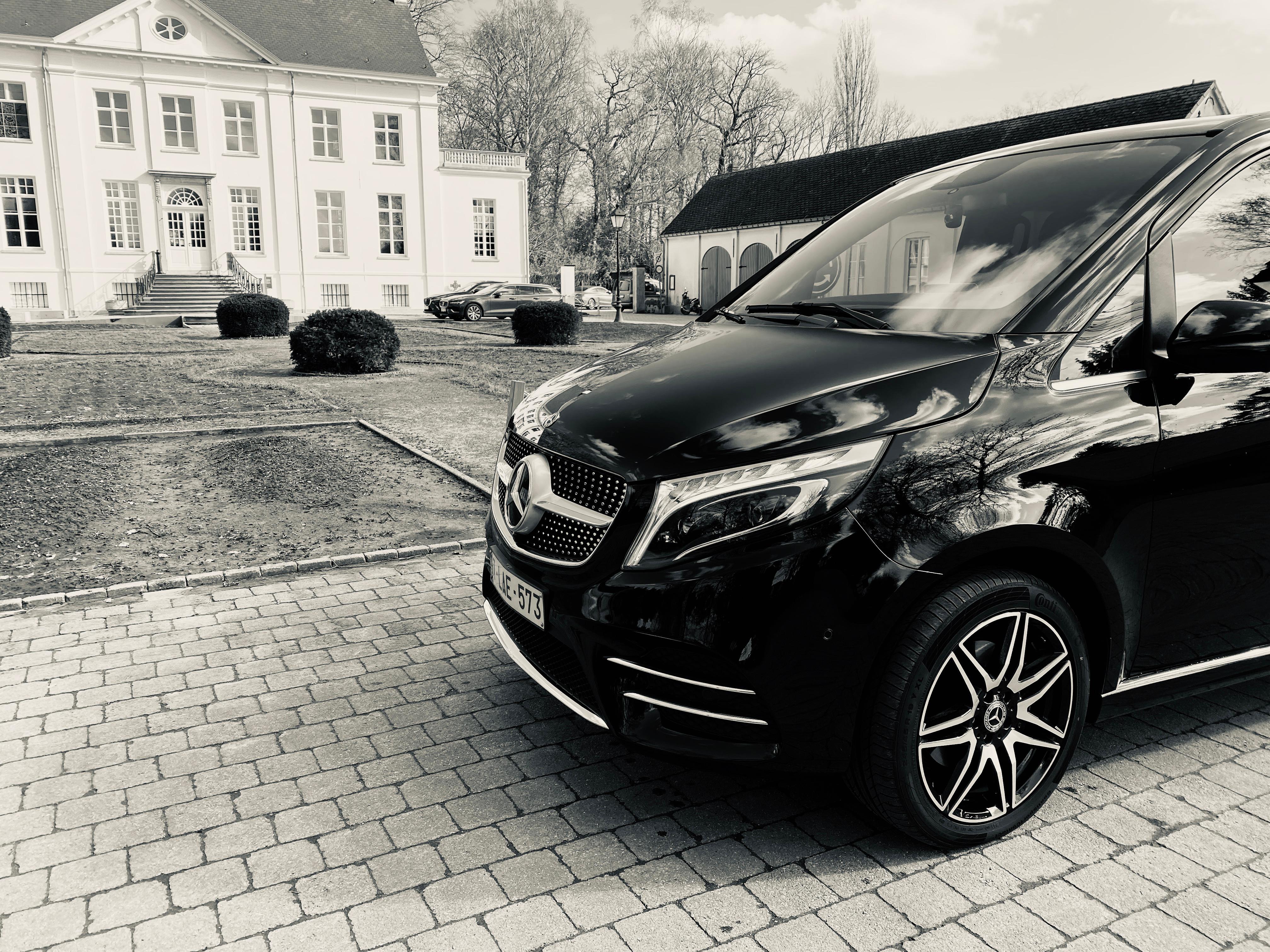 Vooraanzicht van twee schuin geparkeerde wagens: een Mercedes-Benz V-Klasse met chauffeur in maatpak ernaast en een Porsche Taycan, met op de achtergrond een kerk.