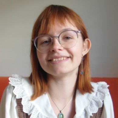 The image shows a person with red hair and glasses, smiling while seated on an orange sofa, wearing a white blouse with lace details, enhancing themes of fashion and style.