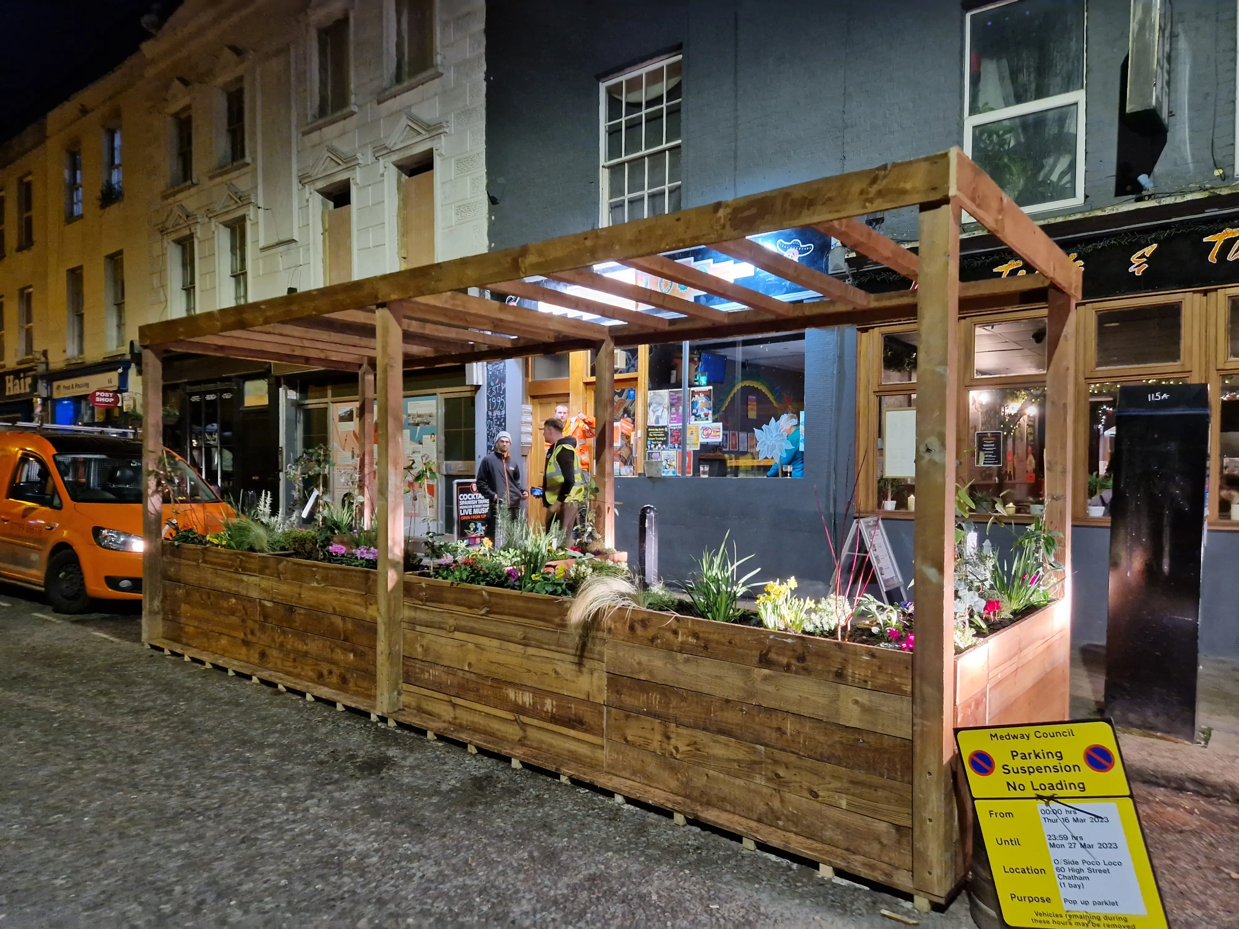 A wooden structure with lights and displays outside a building, creating a lively evening atmosphere on a street.
