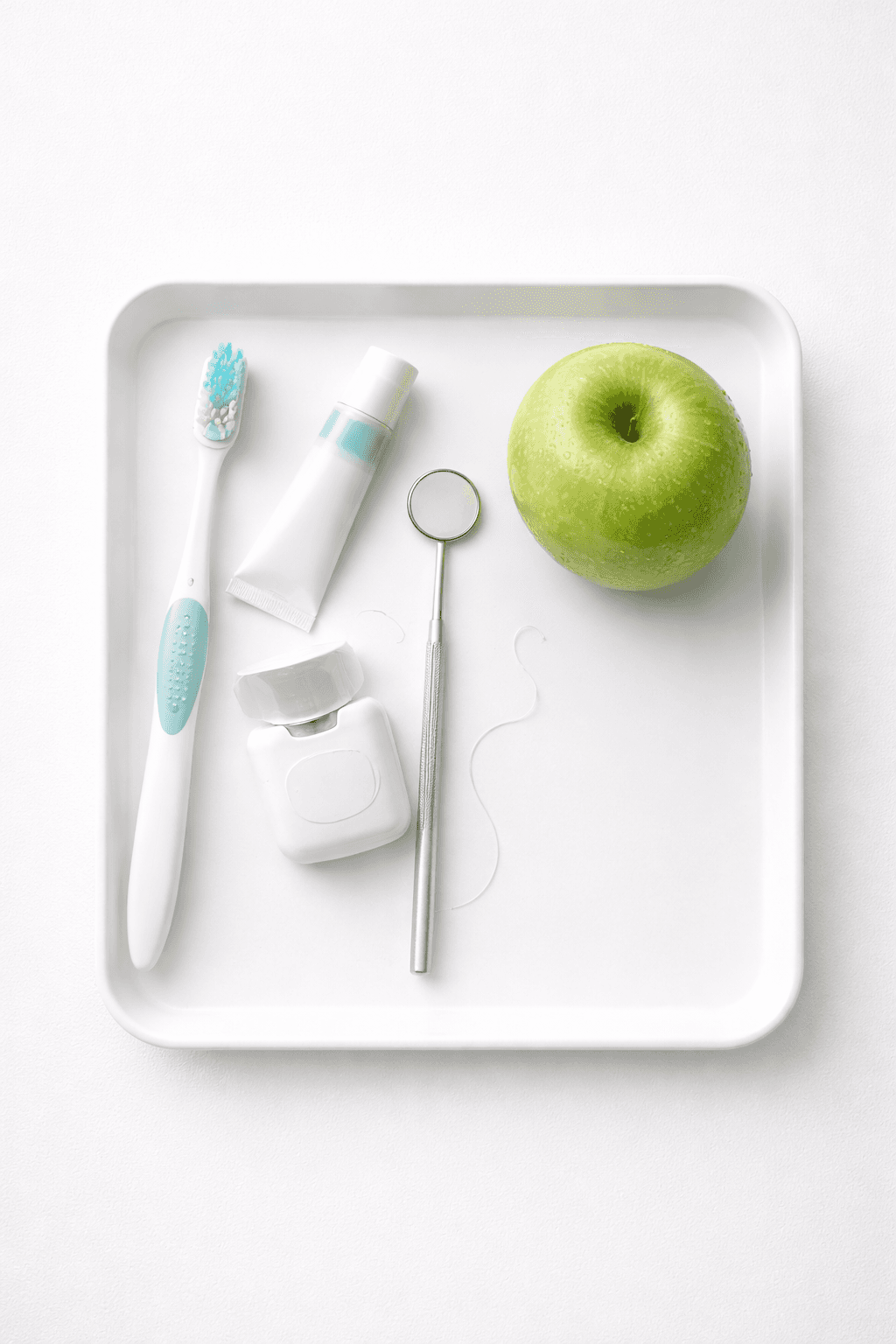 Overhead view of a dental tray containing a toothbrush, dental floss, a green apple, and a small dental mirror on a white background.