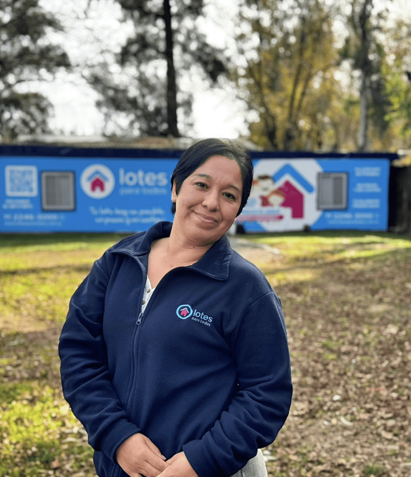 A smiling woman with her arms crossed, standing against a dark green background. She has long, dark hair.