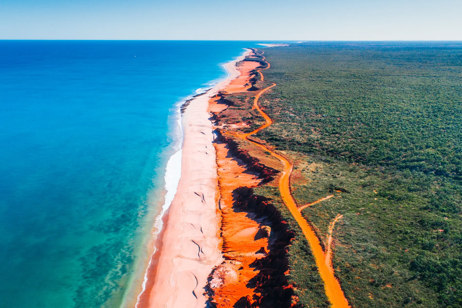 Aerial along a red-earth track beside a long beach, deep blue water on one side and scrub on the other