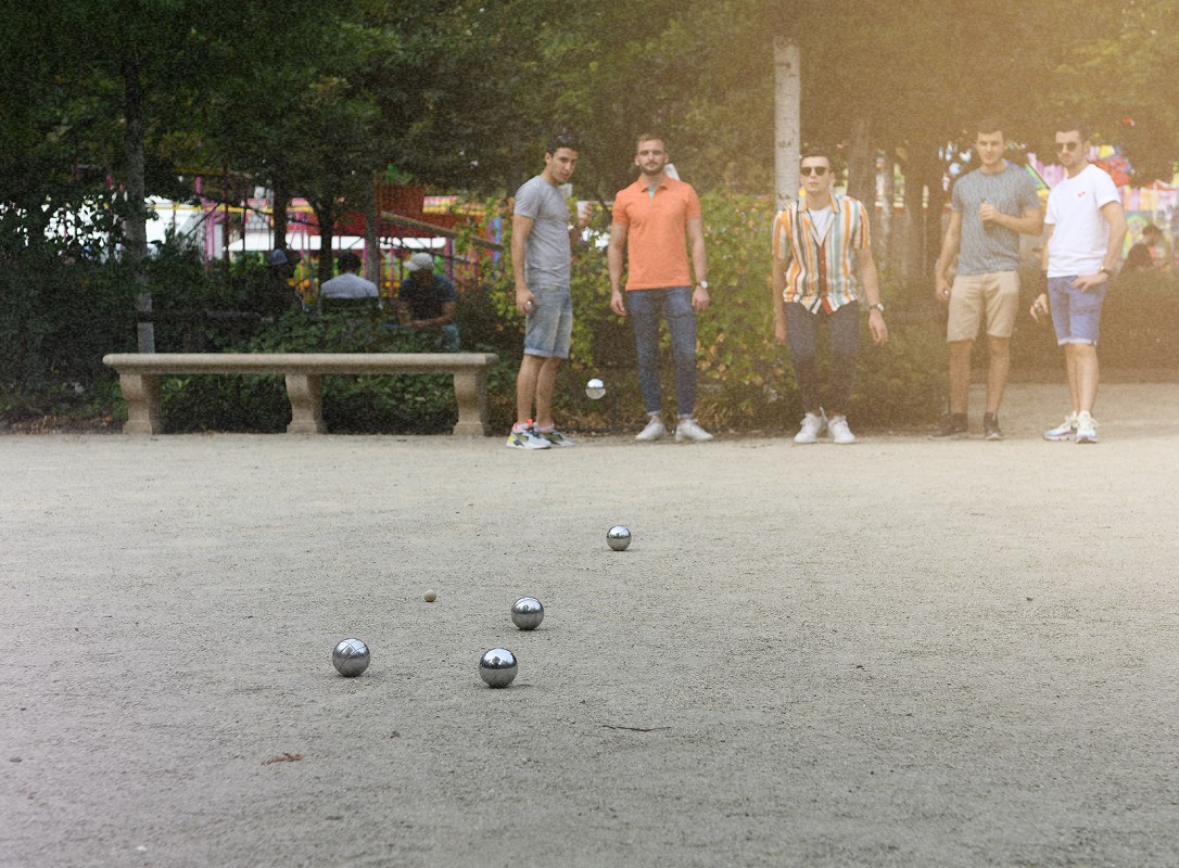 Un groupe de personnes joue à la pétanque dans un parc. Plusieurs boules métalliques sont visibles sur le sol tandis que les joueurs se tiennent en arrière, en attente de leur tour.
