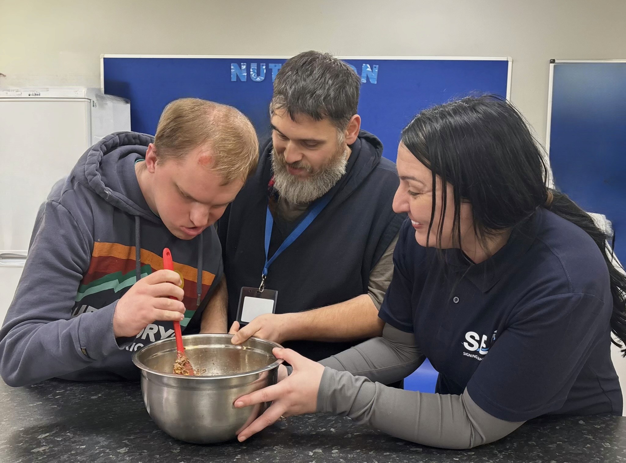  support worker assists a young man with a cooking activity, mixing ingredients in a bowl together at a table