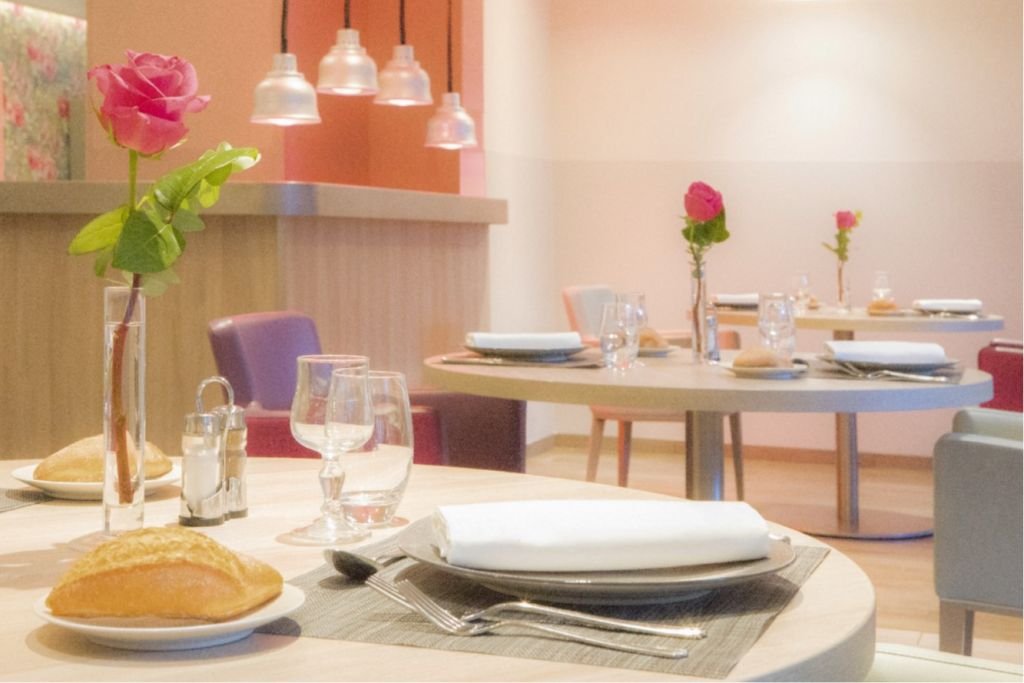 Dining room with round tables set for meals, pink roses in vases, colorful upholstered chairs, and pendant lighting