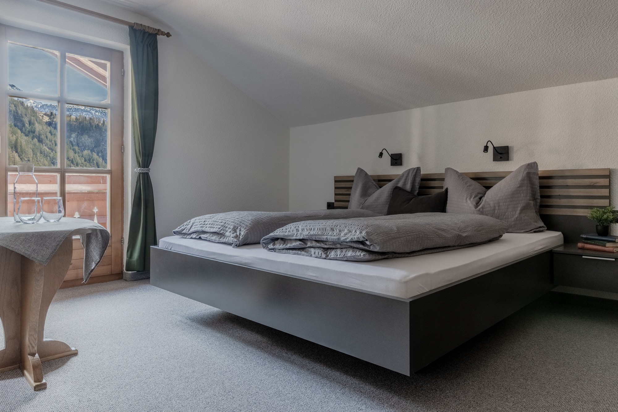 Ein gemütliches, modernes Schlafzimmer verfügt über ein großes Bett mit grauer Bettwäsche, das an einer Dachschräge steht, neben einem Fenster mit malerischem Bergblick, ergänzt durch einen kleinen Holztisch mit zwei Gläsern.
