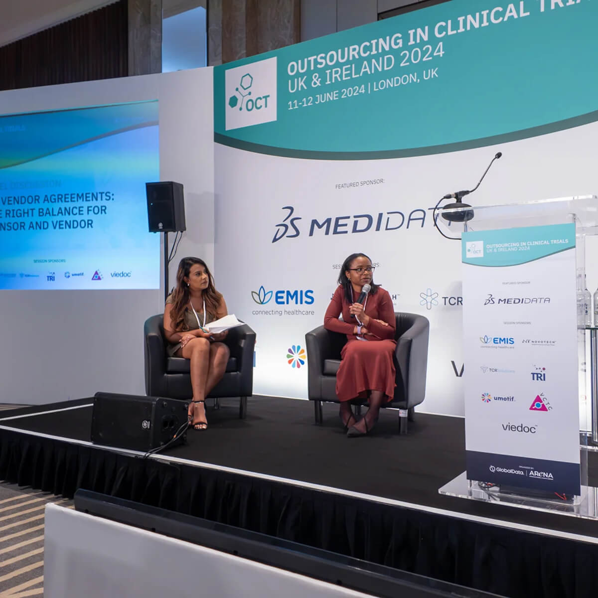 Two people sat on a stage speaking at an event. A stage backdrop, lectern and slide screen is shown too.