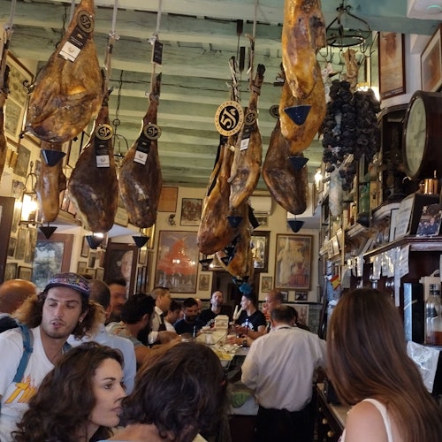 A busy tapas bar with customers crowding the counter, walls adorned with framed pictures, and cured hams hanging from the ceiling.