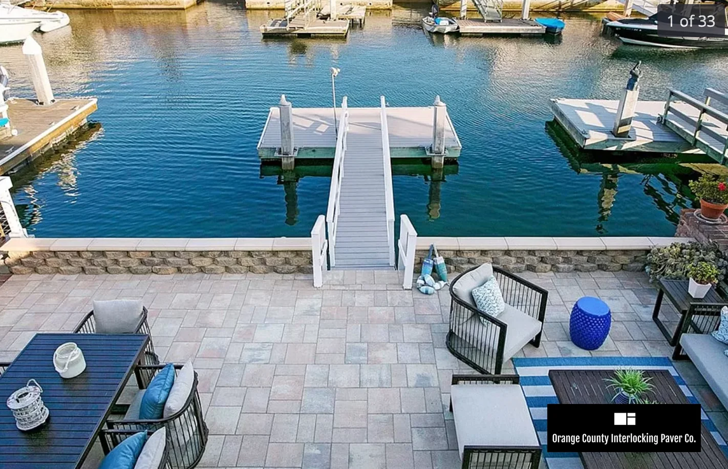 Waterfront paver patio with outdoor chairs, tables and a walkway leading to a dock.